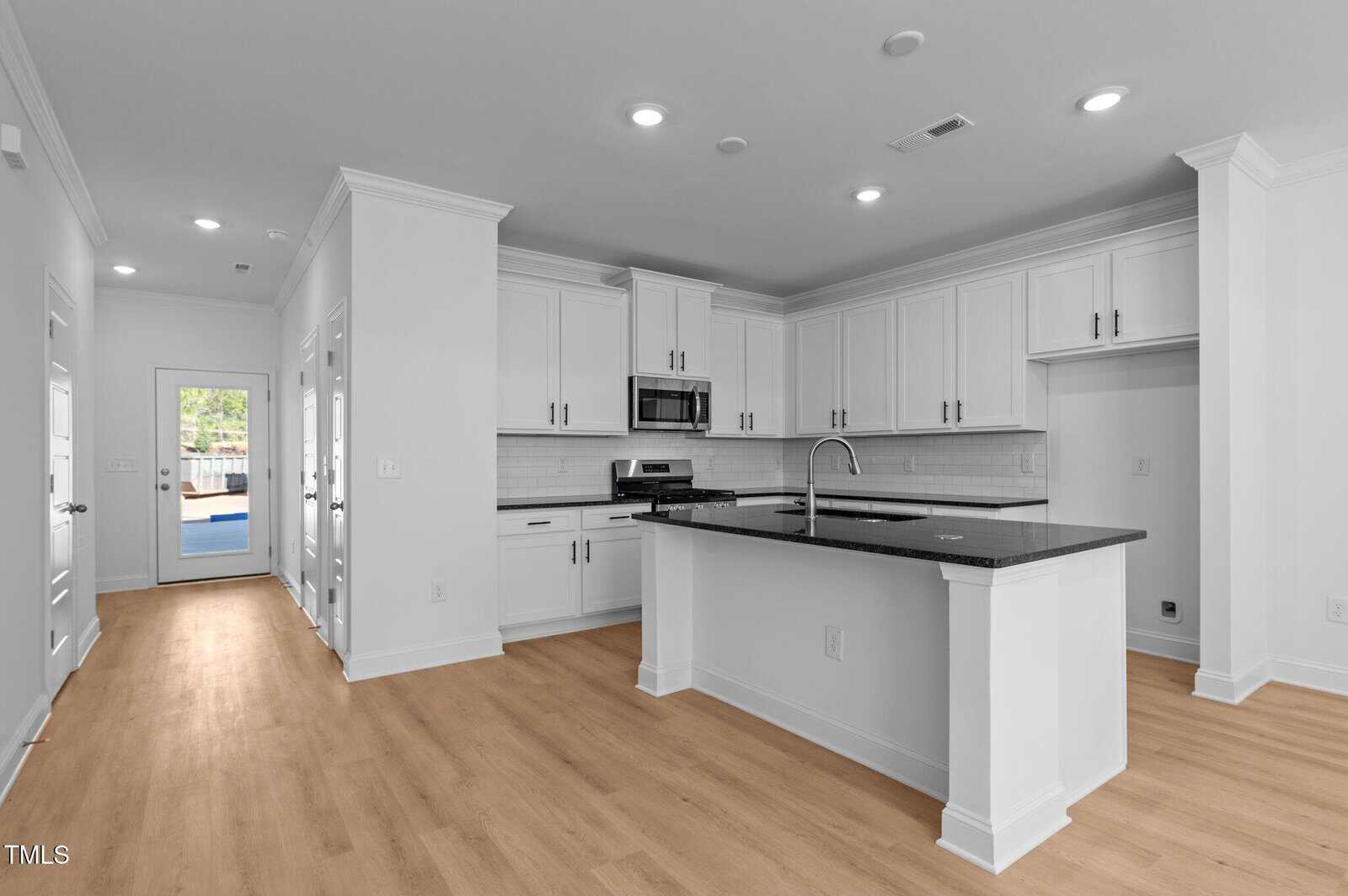 Modern kitchen with white cabinets, black countertops, stainless steel appliances, an island with a sink, light wood flooring, and bright overhead lighting; a door and windows are visible in the background.