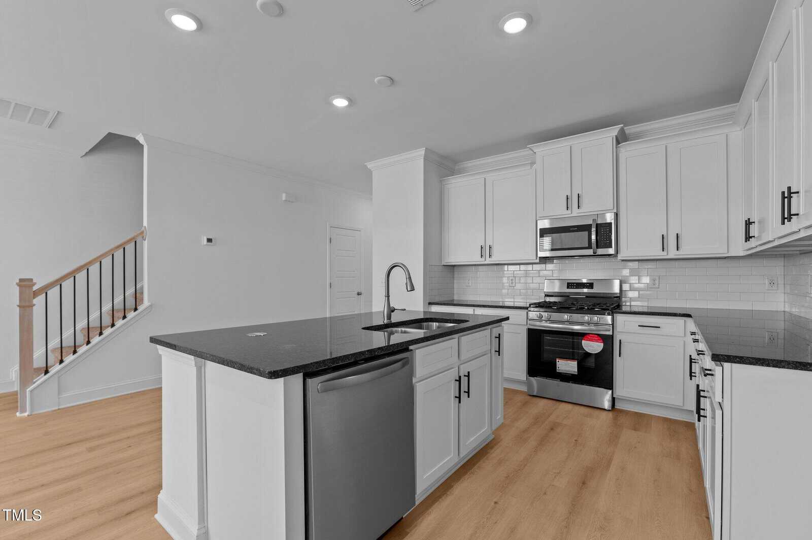 Modern kitchen with white cabinets, black countertops, stainless steel appliances, an island with a sink, light wood flooring, and a staircase visible to the left.