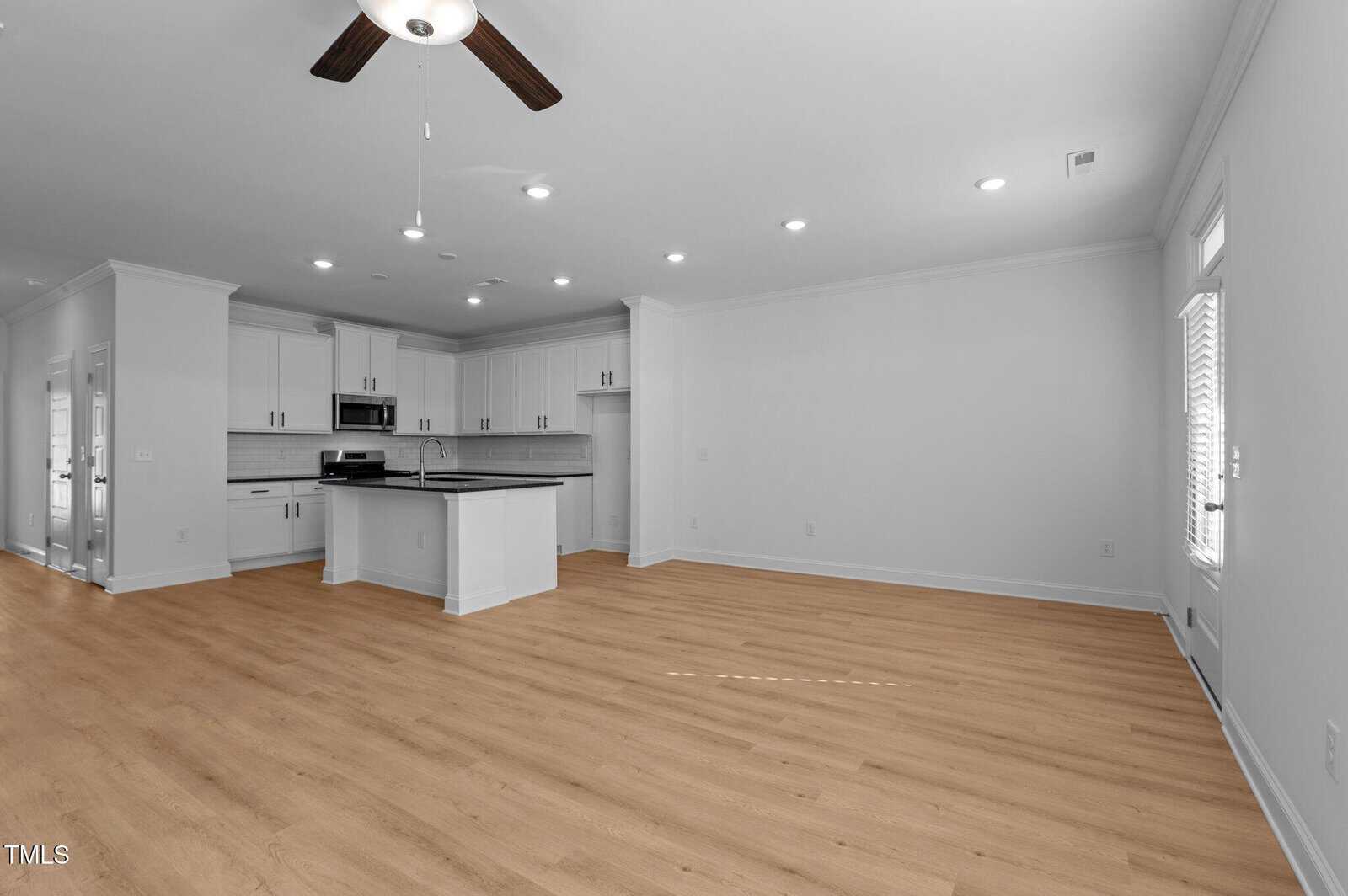 Spacious, modern open-plan kitchen and living area with light wood flooring, white cabinets, stainless steel appliances, and large windows providing natural light. Ceiling fan and recessed lighting are visible.