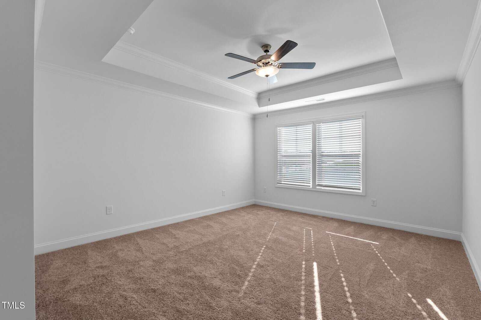A bright, empty room with beige carpet, white walls, a tray ceiling, a ceiling fan with lights, and two large windows letting in natural sunlight.
