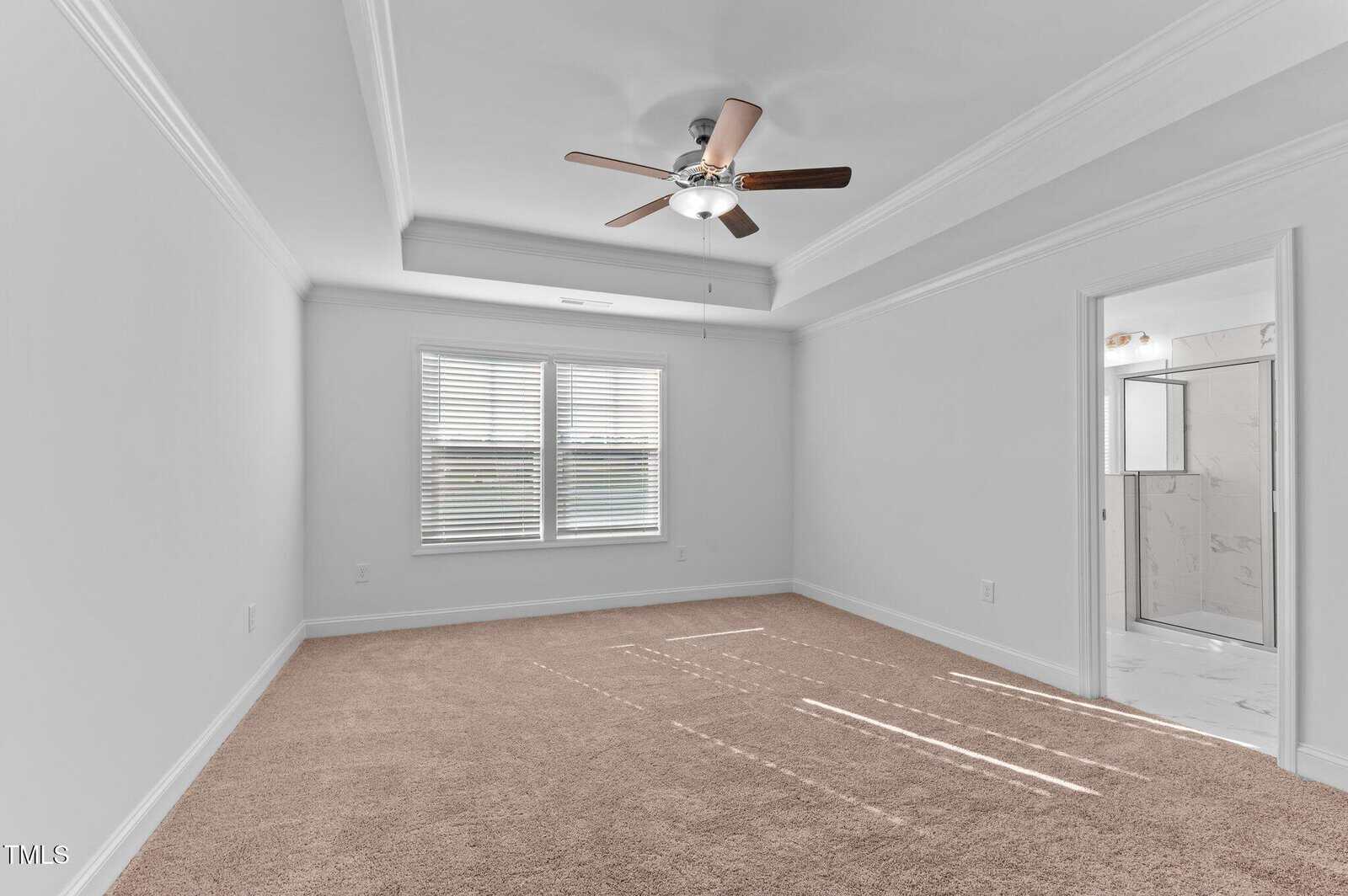 Empty bedroom with beige carpet, white walls, a ceiling fan with wooden blades, two windows with blinds, and a doorway leading to a bathroom with a glass shower enclosure.