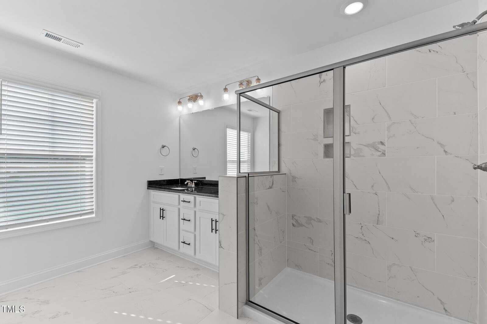 Modern bathroom with white marble tile flooring and walls, a glass-enclosed shower, a vanity with a black countertop and double sinks, two mirrors, light fixtures above, and a window with white blinds.