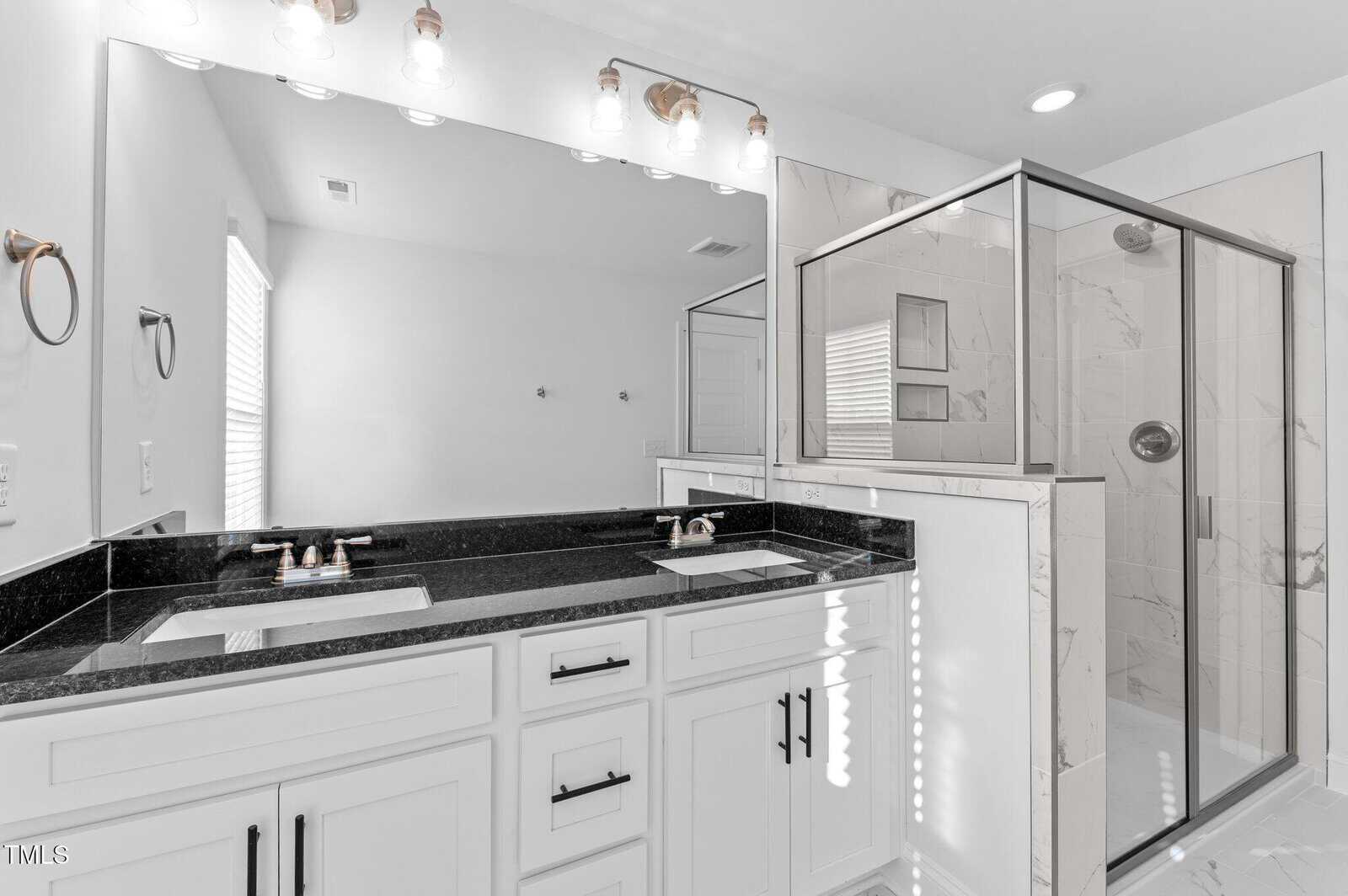 Modern bathroom with double sinks, black countertops, white cabinets, and a large glass-enclosed shower with marble tile. Above the sinks are three light fixtures, and a mirror runs the length of the vanity.