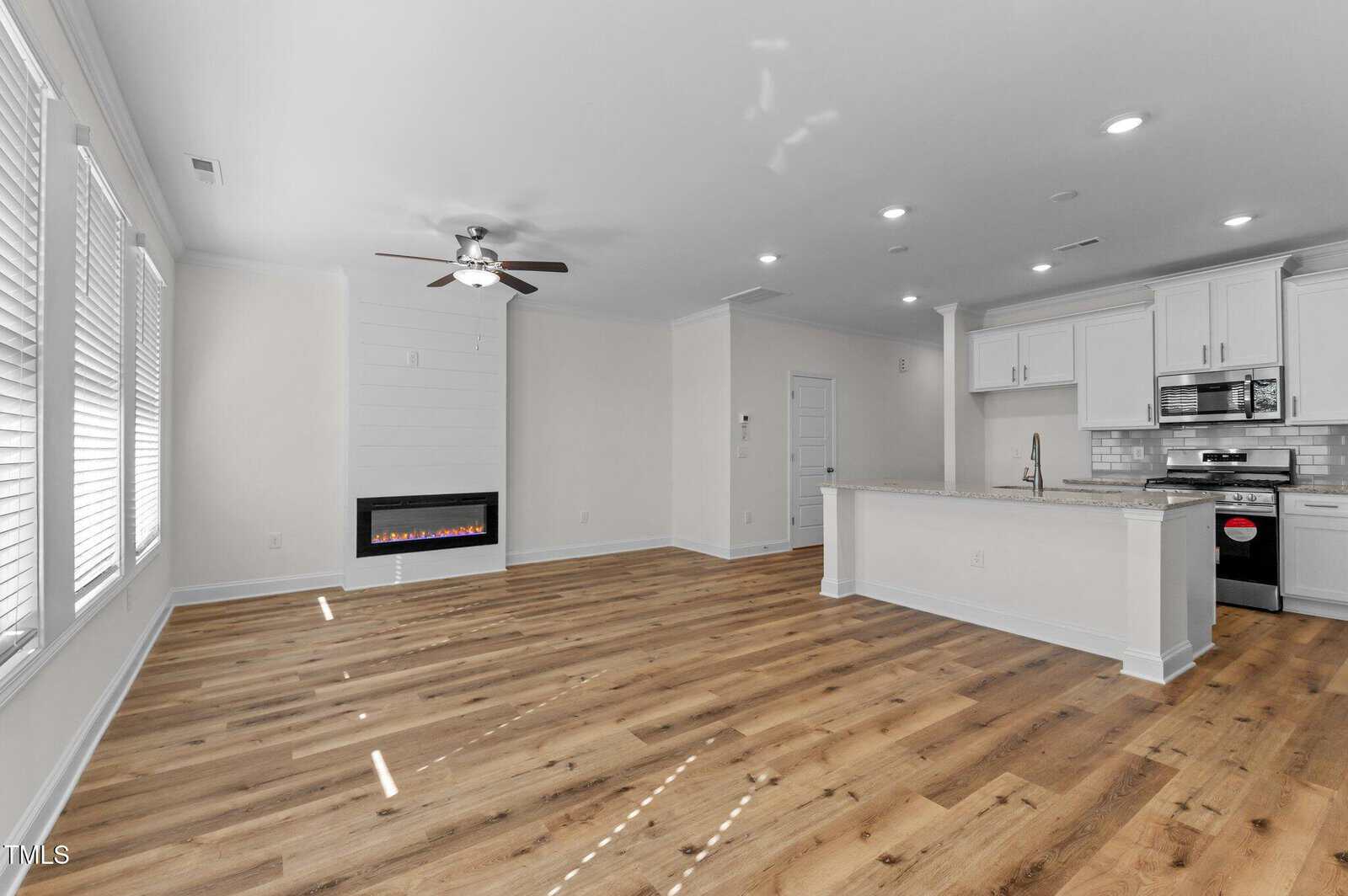 A modern, unfurnished open-concept living area with light wood floors, white walls, a ceiling fan, electric fireplace, and a kitchen with white cabinets, subway tile backsplash, and stainless steel appliances.