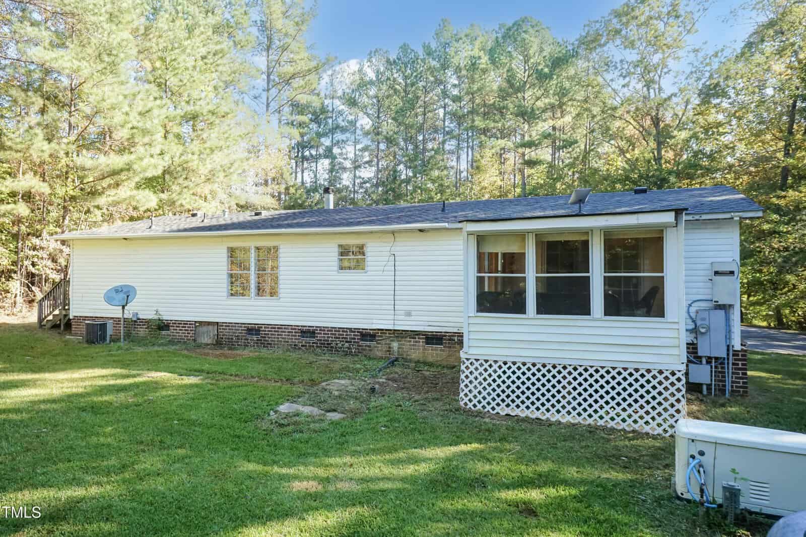 A single-story white house with a screened porch is surrounded by tall trees. The house has a dish antenna and brick foundation. The lawn is green and spacious, extending to the edge of the woods.