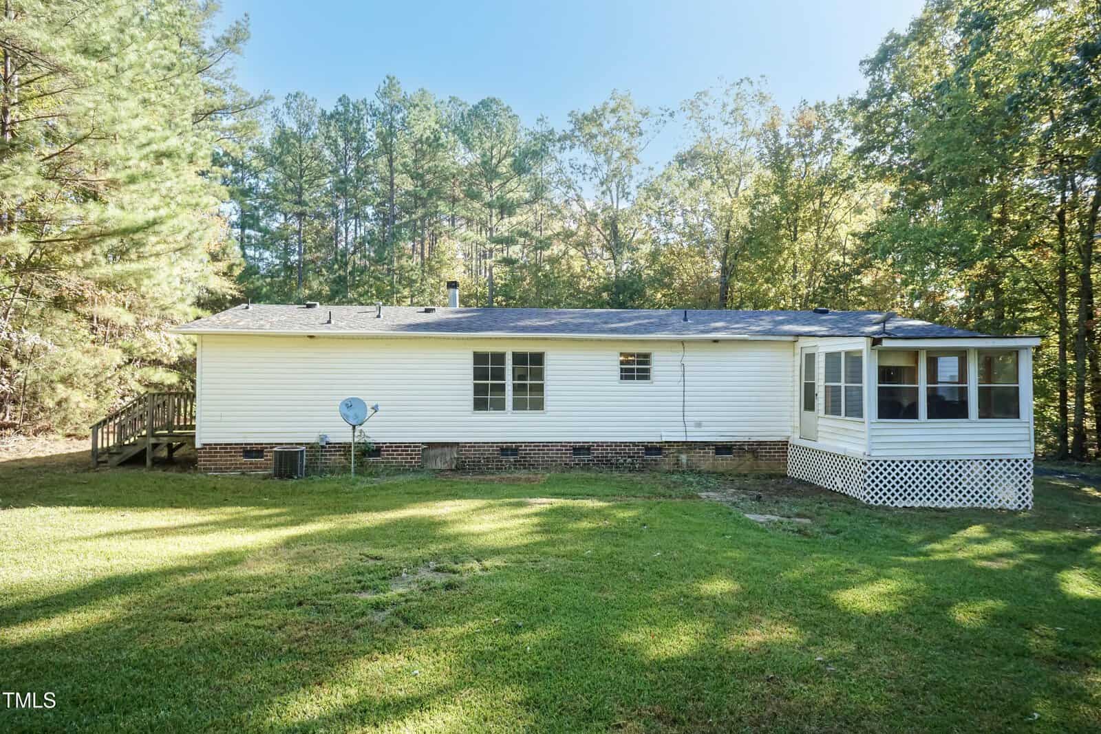 A single-story white house with a screened porch at the back, situated on a grassy lawn. Surrounded by trees, the house has a satellite dish and a sloping roof. The siding appears to be vinyl, and there is a wooden ramp on one side.
