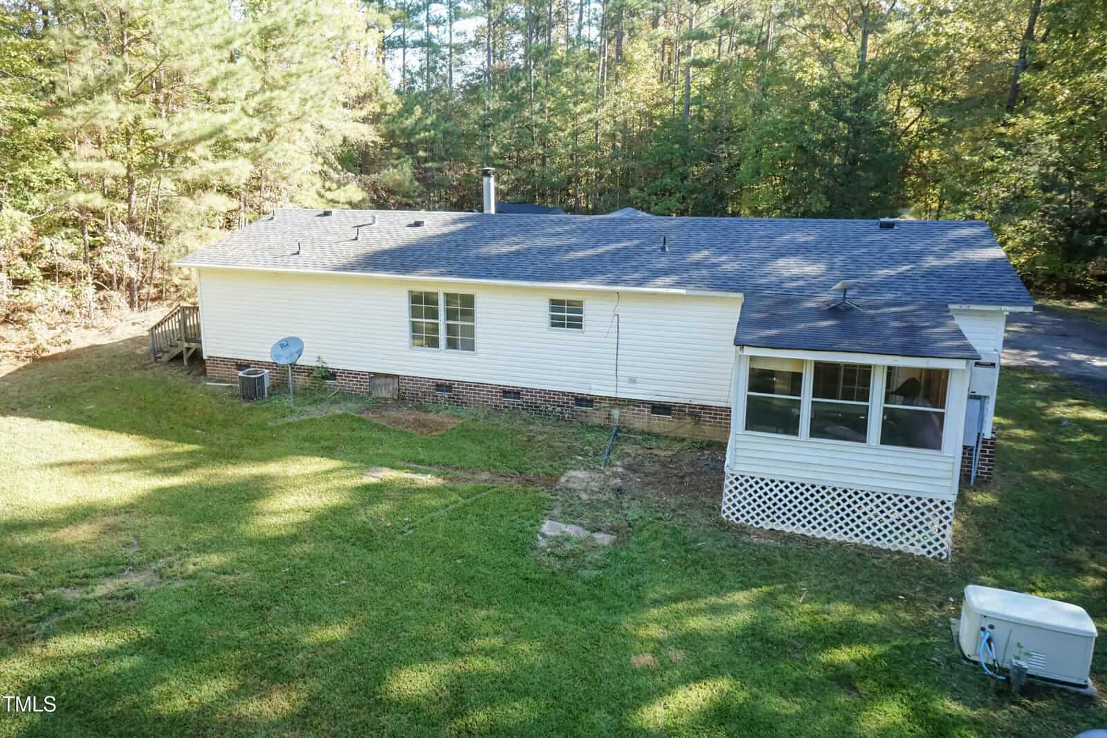 A single-story white house with a dark roof is surrounded by a grassy yard and trees. It features a screened porch, a satellite dish, and an air conditioning unit. The backyard is partially shaded by tall trees in the background.
