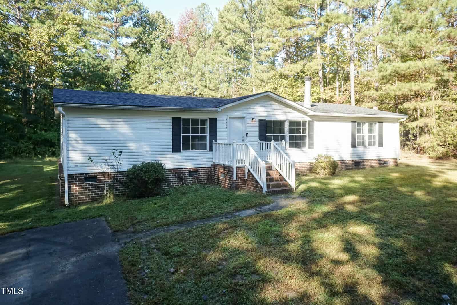 Located at 7589 Sam Hall Road in Oxford, this single-story white house boasts dark shutters and a charming porch. It's enveloped by lush grass and trees, with a short driveway leading invitingly to the entrance.