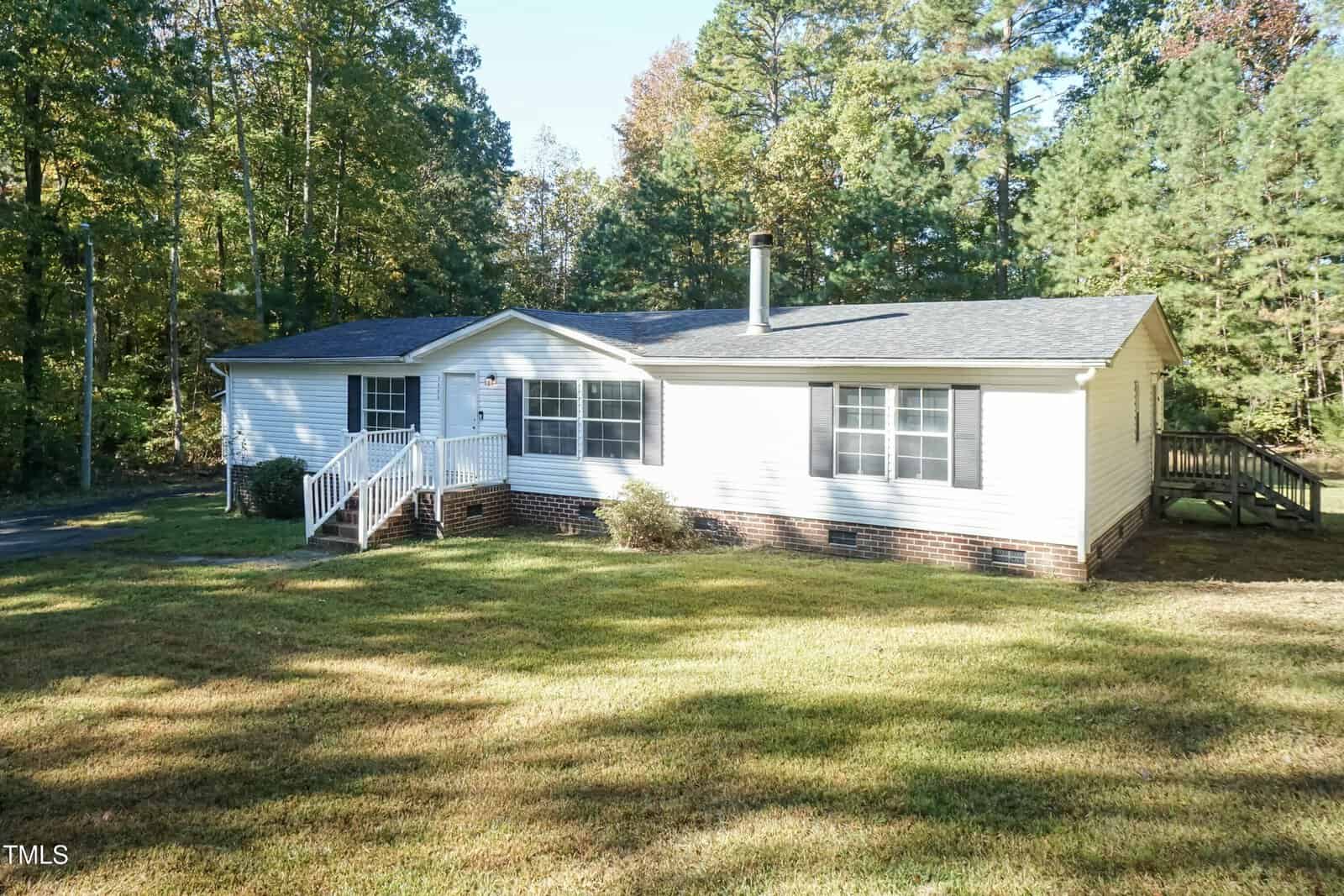 A white single-story house with a brick foundation, surrounded by trees. It has a small porch with steps, a dark roof, and a lawn in the foreground. The driveway is partially visible on the left. The scene is in a wooded area on a sunny day.