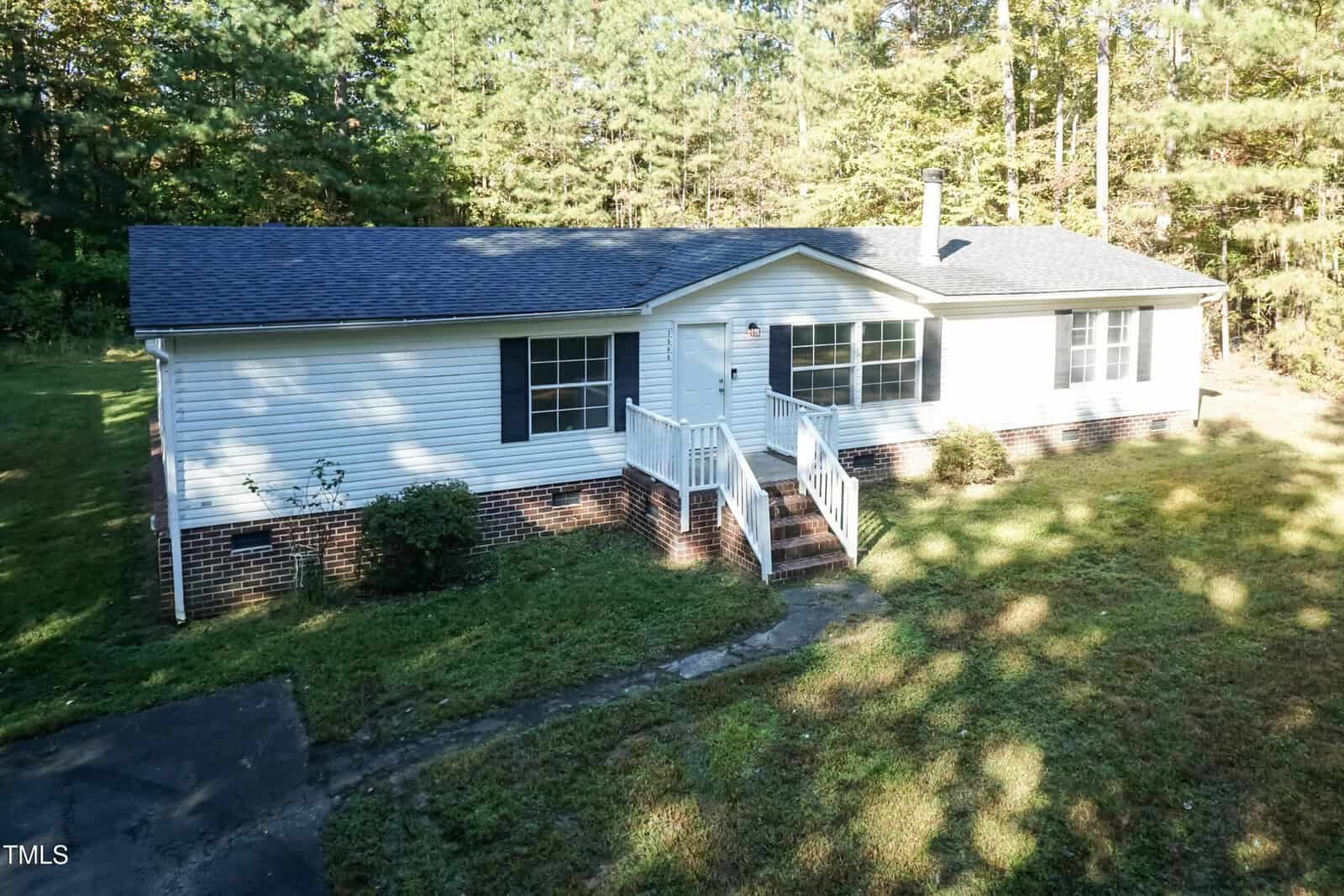A single-story house with white siding and a dark shingled roof is surrounded by grass and trees. The front features a small porch with steps leading down to a path. There is a paved area on the left side.