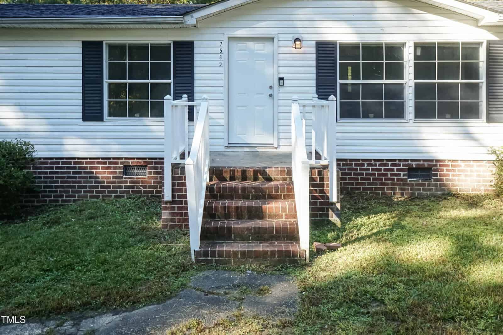 A small white house with a brick foundation, featuring a central white door flanked by two windows with black shutters. A set of brick steps leads up to the entrance, and the front yard is partially covered with grass.