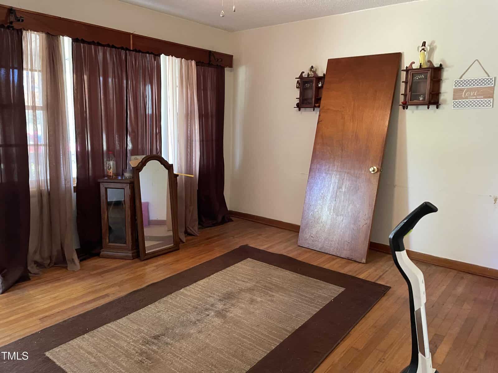 An empty room with a wooden floor, a tilted door leaning against the wall, dark curtains, a mirror, and an exercise bike. Shelves and a sign are mounted on the wall. A large rug covers part of the floor.