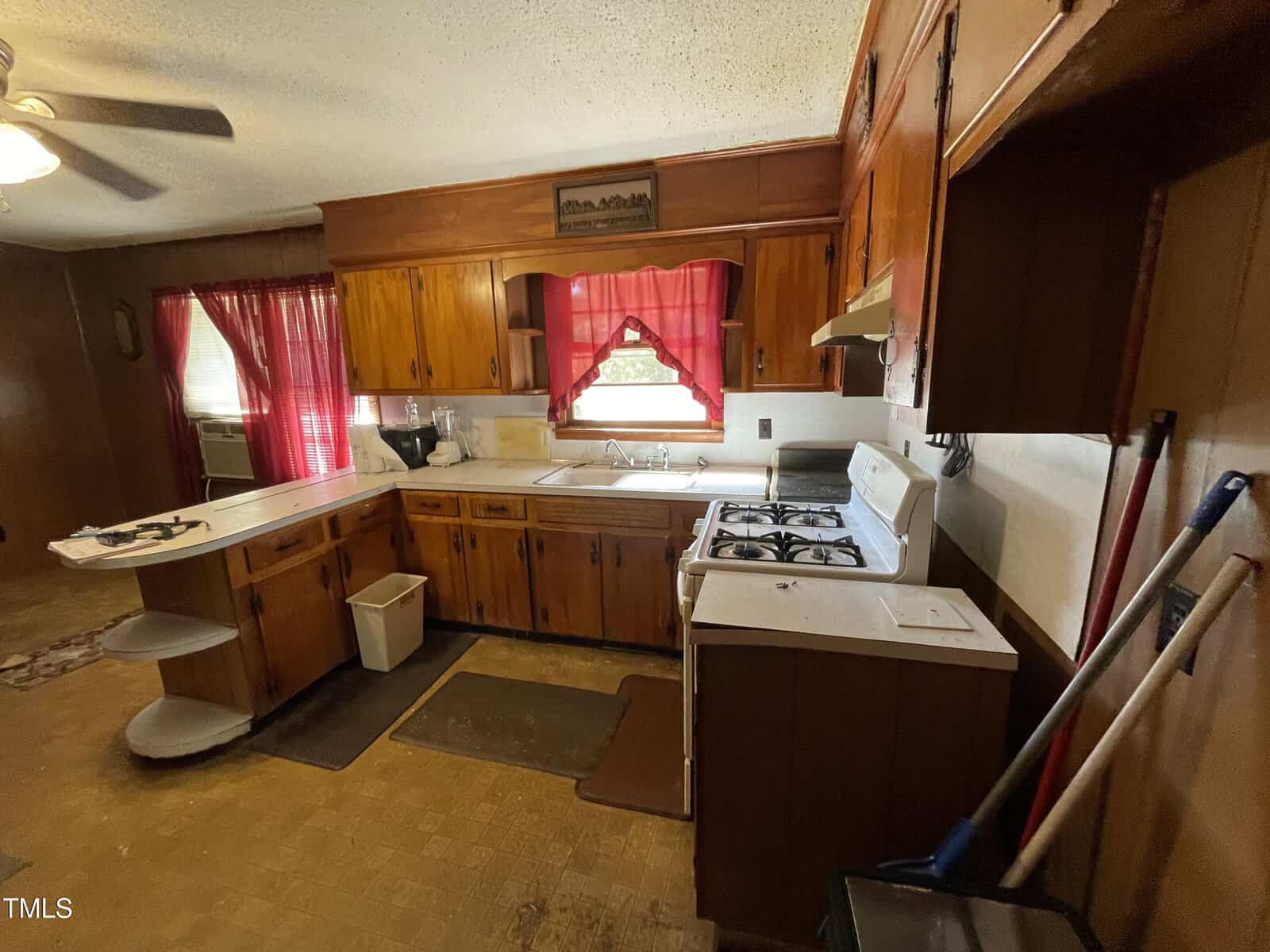 A vintage-style kitchen with wooden cabinets, a white gas stove, and a dual sink. Red curtains adorn the window above the sink. A ceiling fan hangs above, and cleaning tools are stored in the corner.