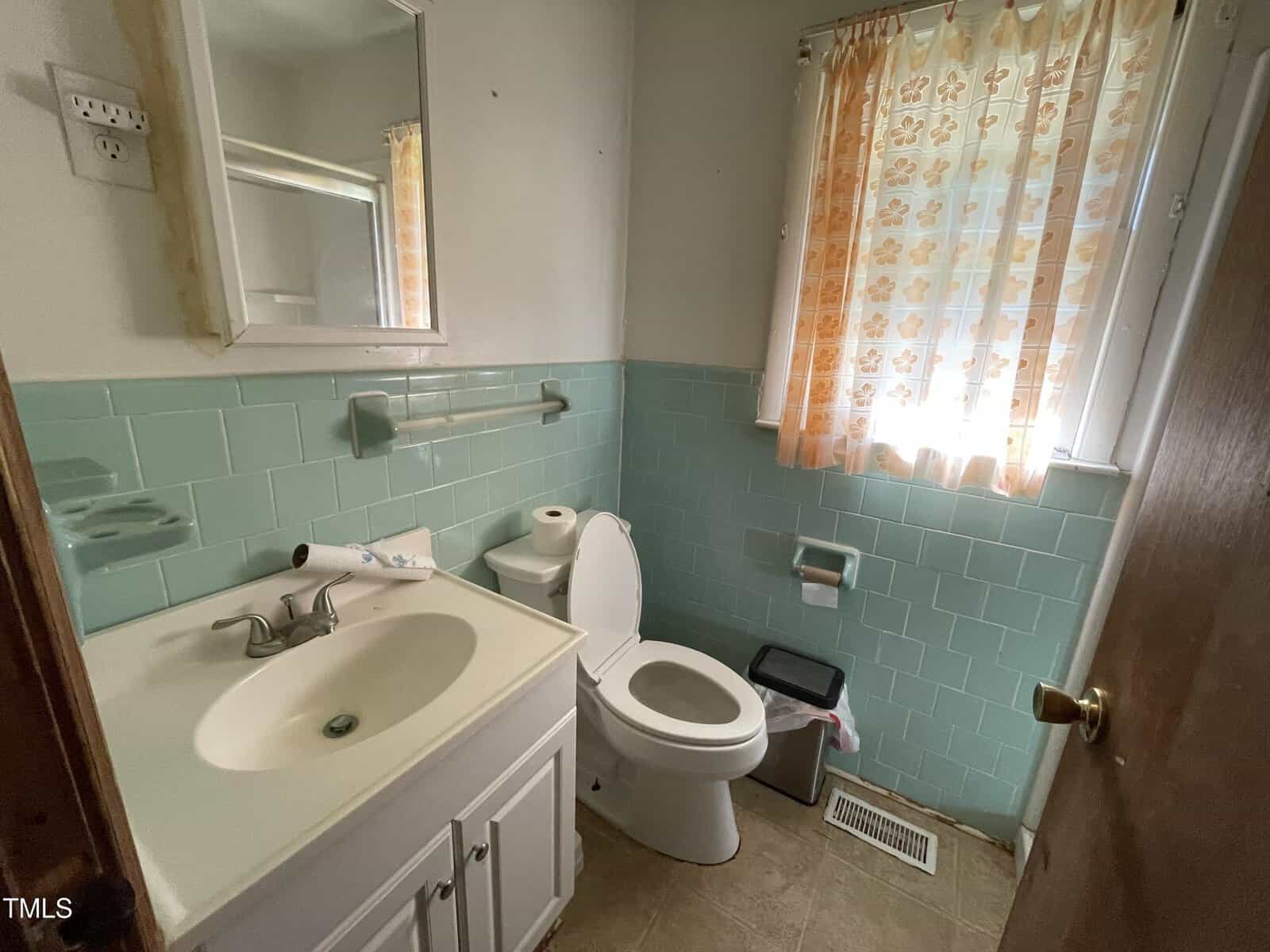 A small bathroom with mint green tile walls, a white toilet, and a white sink with a vanity. A floral-patterned curtain covers a window, and there is a wooden door slightly ajar. The floor is light brown tile.