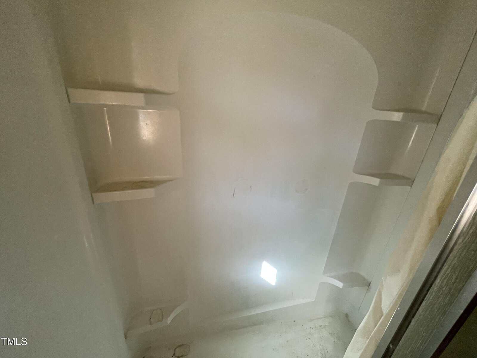 A white shower stall with curved walls and built-in shelves on both sides. Sunlight casts a small bright rectangle on the back wall. The surface appears clean and unused.