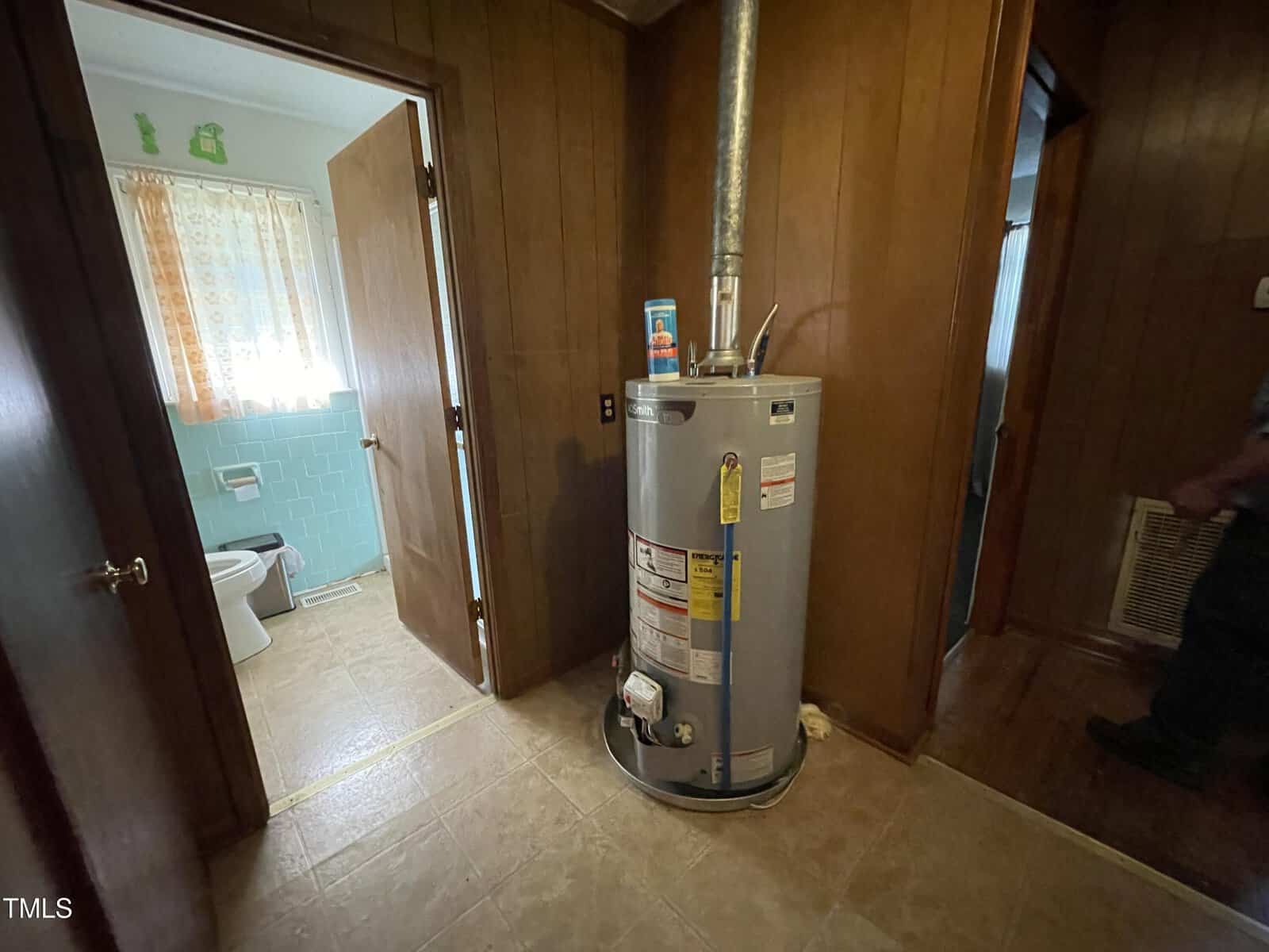 A home interior with wooden paneled walls features a water heater in a small hallway space. An open door on the left reveals a bathroom with light blue tiles and floral-patterned curtains. A can and bottle are placed on top of the heater.
