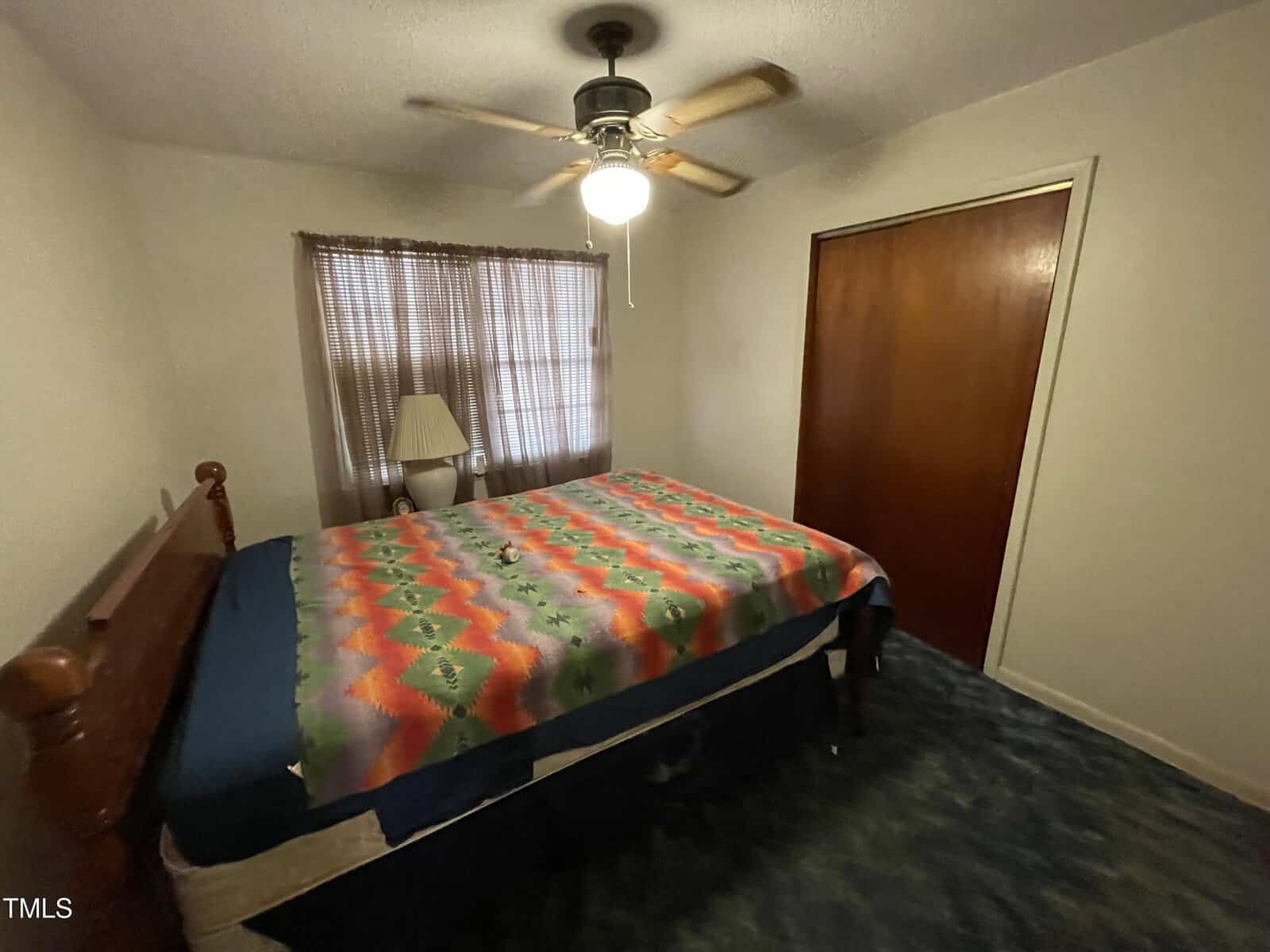 A small bedroom with a colorful patterned bedspread on the bed, a wooden headboard, a ceiling fan, and a window with sheer curtains. There's a floor lamp beside the bed and a closed closet door on the right wall.