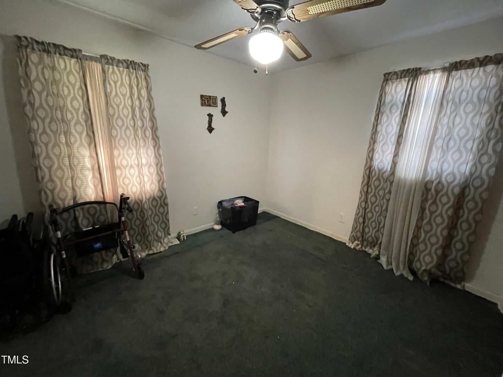 A dimly lit room with green carpet, a ceiling fan, two windows with patterned curtains, and a few wall decorations. A walker and a plastic storage bin are against the walls, and the room appears sparsely furnished.