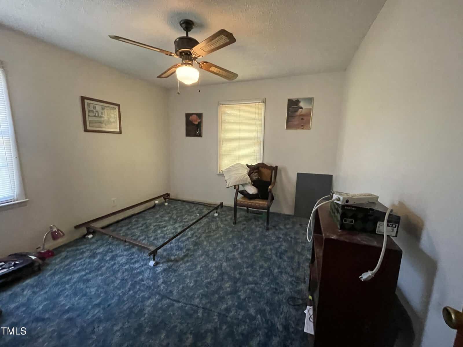 A small, empty bedroom with a blue carpet, a ceiling fan, a single chair with clothes on it, and a bed frame without a mattress. The room has a window, a few pictures on the walls, and some equipment on a dresser.