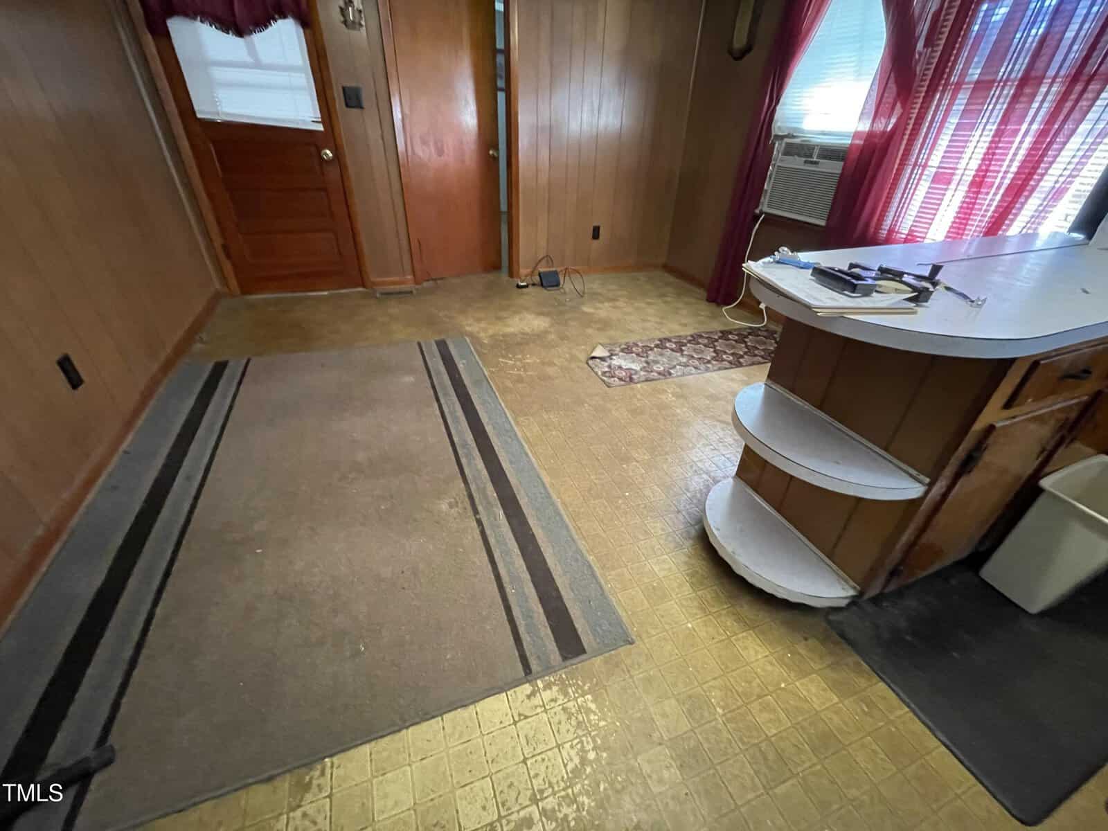 A small room with wood-paneled walls, a brown carpet, and linoleum flooring. A door is partially visible along with a window with curtains and an air conditioner. A curved countertop with shelves is on the right. A rug and a trash can are also visible.
