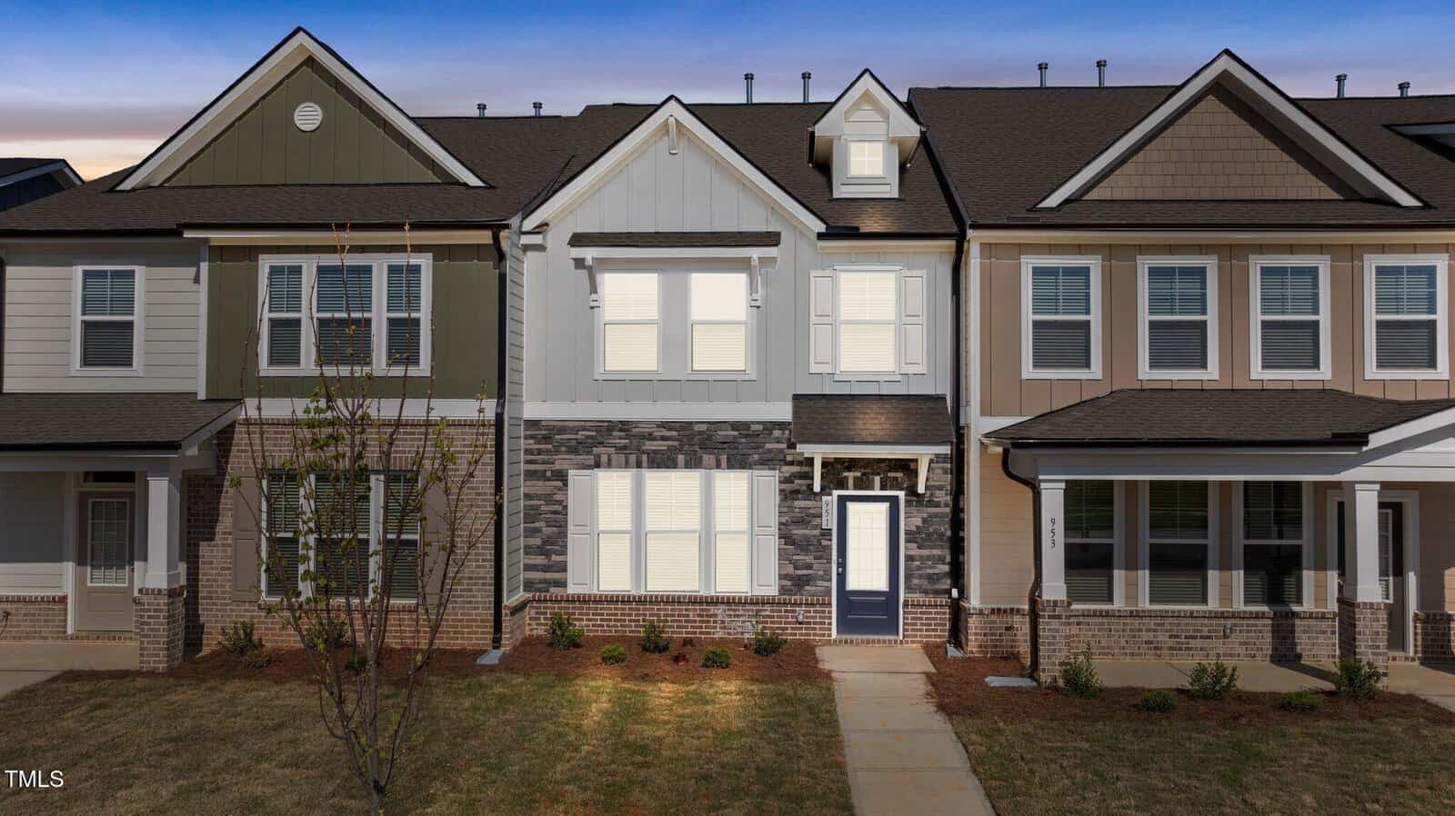 A two-story townhouse with a mix of white, tan, and sage green paneling, dark stone accents, white trim, and a blue front door. Small shrubs and a young tree are planted in the front yard at dusk.