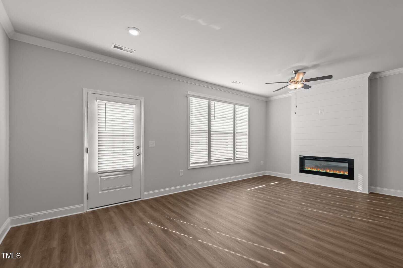 Bright, modern living room with light gray walls, wood flooring, large windows, a door with blinds, and a wall-mounted electric fireplace below a ceiling fan. Natural light fills the space.