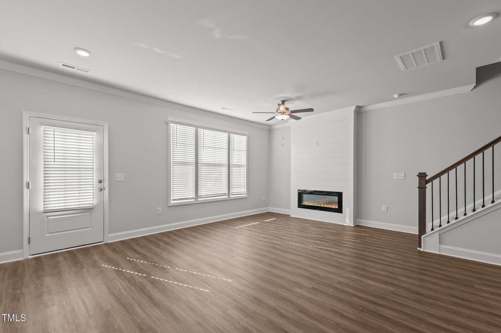 Spacious living room with light wood flooring, large windows, a ceiling fan, and a modern electric fireplace beside a staircase with a dark wooden handrail.