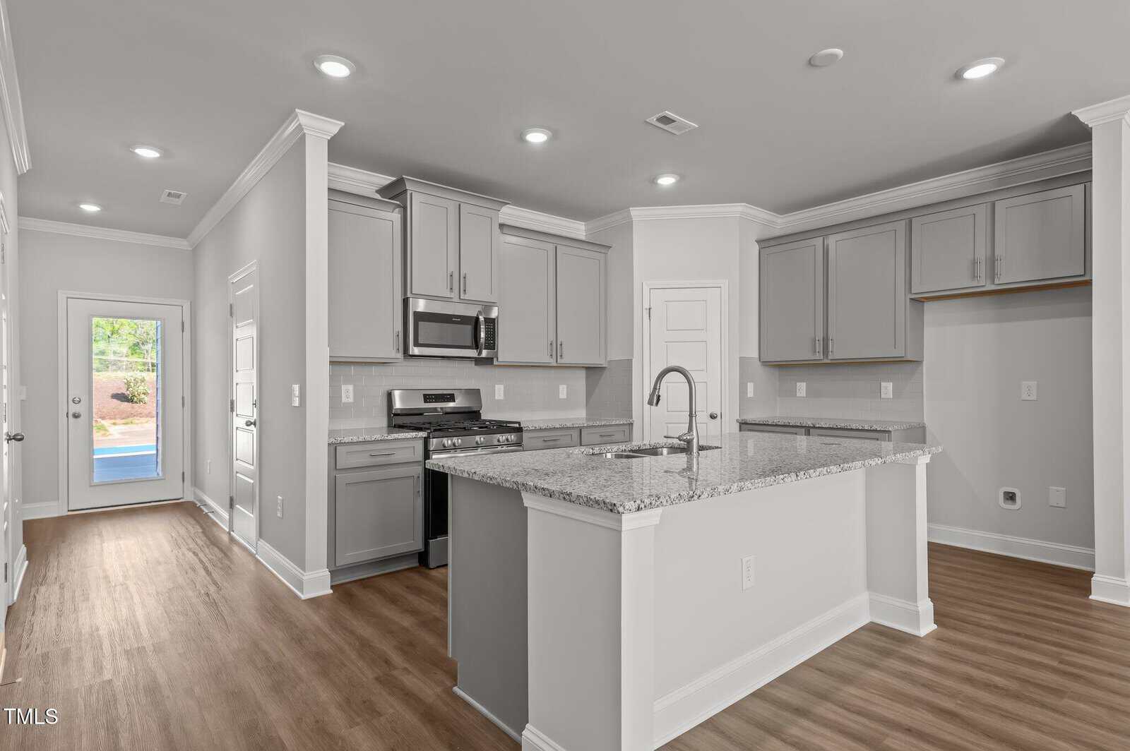 Modern kitchen with gray cabinets, stainless steel appliances, granite countertops, a large center island with a sink, and wood flooring. A door at the back leads outside, letting in natural light.