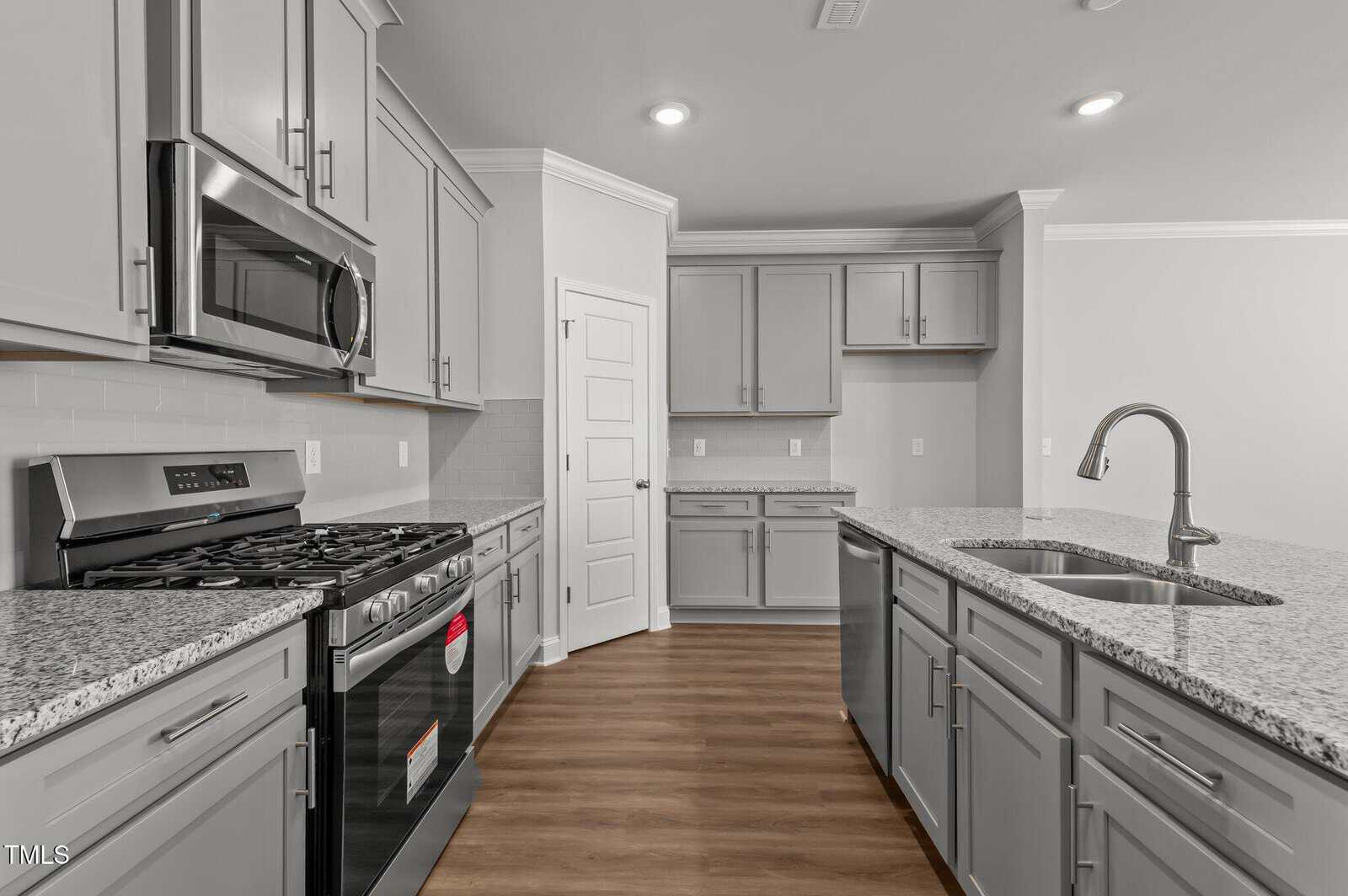 Modern kitchen with gray cabinets, stainless steel appliances, a gas stove, built-in microwave, granite countertops, a large island with a sink, and wood flooring. White door and light gray walls in the background.