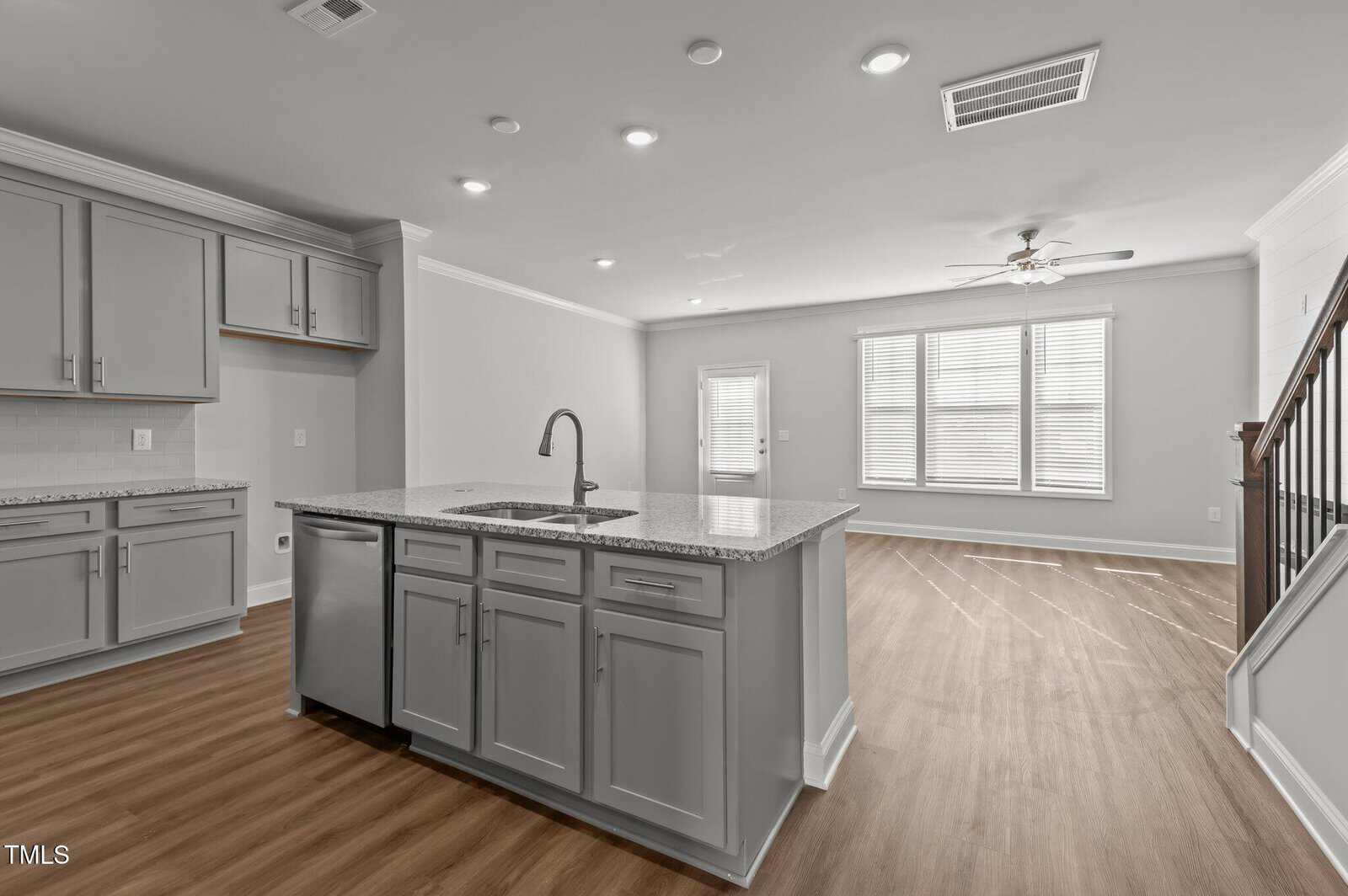 Modern, open-concept kitchen and living area with gray cabinets, granite island, stainless steel faucet, light wood floors, large windows, and a staircase to the right. Room is bright with recessed lighting and neutral walls.