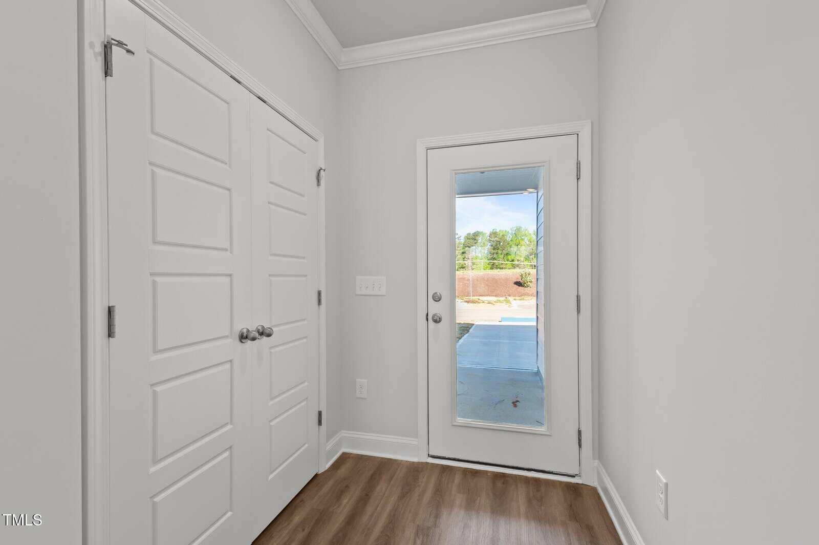 A hallway with light gray walls, white trim, double closet doors on the left, wood-look flooring, and a glass door leading outside to a covered patio and fenced yard.