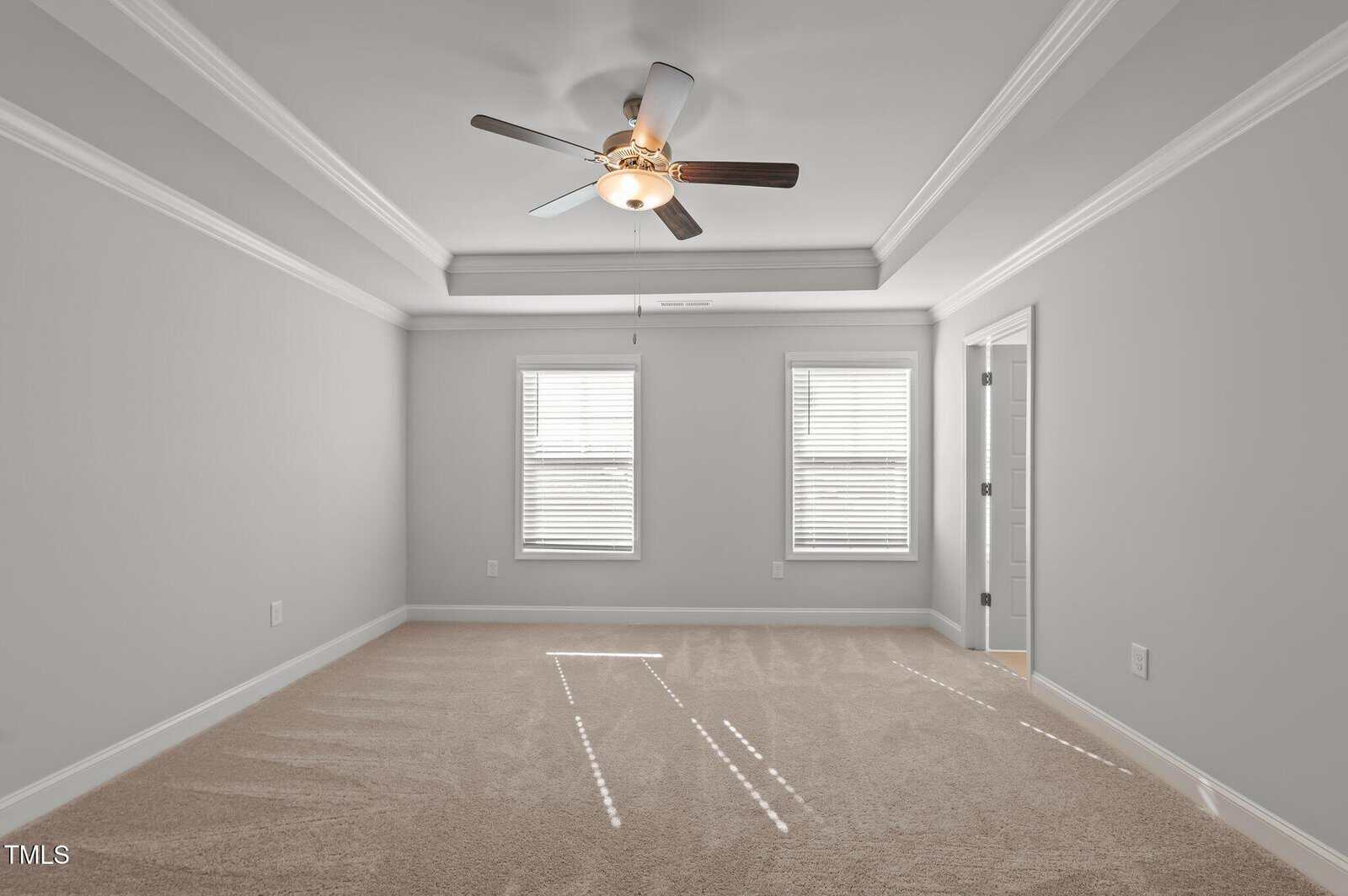 Empty bedroom with beige carpet, light gray walls, two windows with blinds, a ceiling fan with light, and a doorway to the right leading to another room.