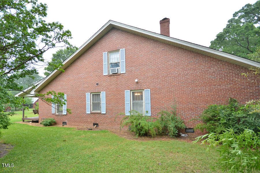 A one-story house with a red brick exterior and light blue shutters. The side yard is grassy with trees and shrubs, and a small window unit air conditioner is visible. A chimney is seen on the roof.
