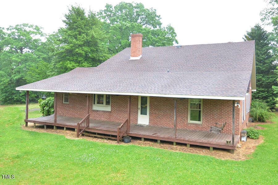 A brick house with a large sloped roof and a spacious wooden porch. The porch has steps leading down to a grassy yard. There are two doors and several windows along the front of the house, surrounded by trees and greenery.