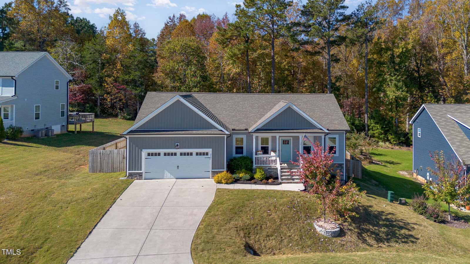 Aerial view of a single-story gray house with a front porch, two-car garage, and a sloped driveway, surrounded by a green lawn, trees with autumn foliage, and neighboring homes.