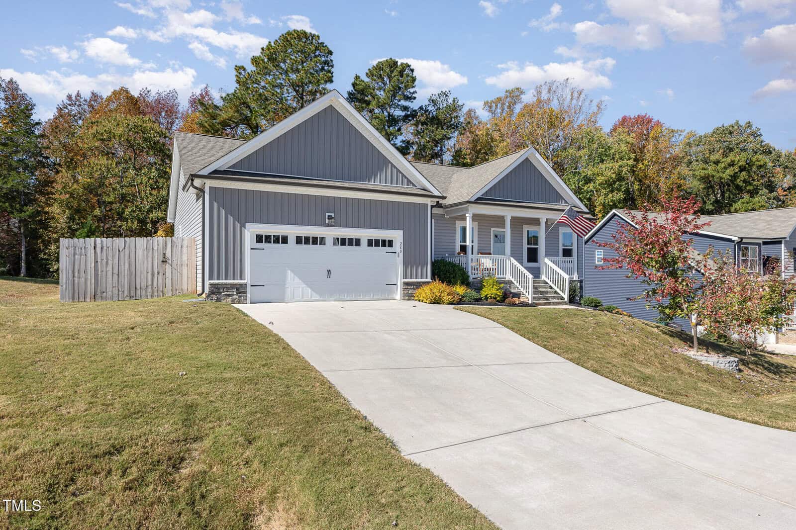A gray single-story house with a two-car garage, front porch, and American flag, sits on a sloped lawn with a concrete driveway. Trees with autumn foliage are in the background under a partly cloudy sky.