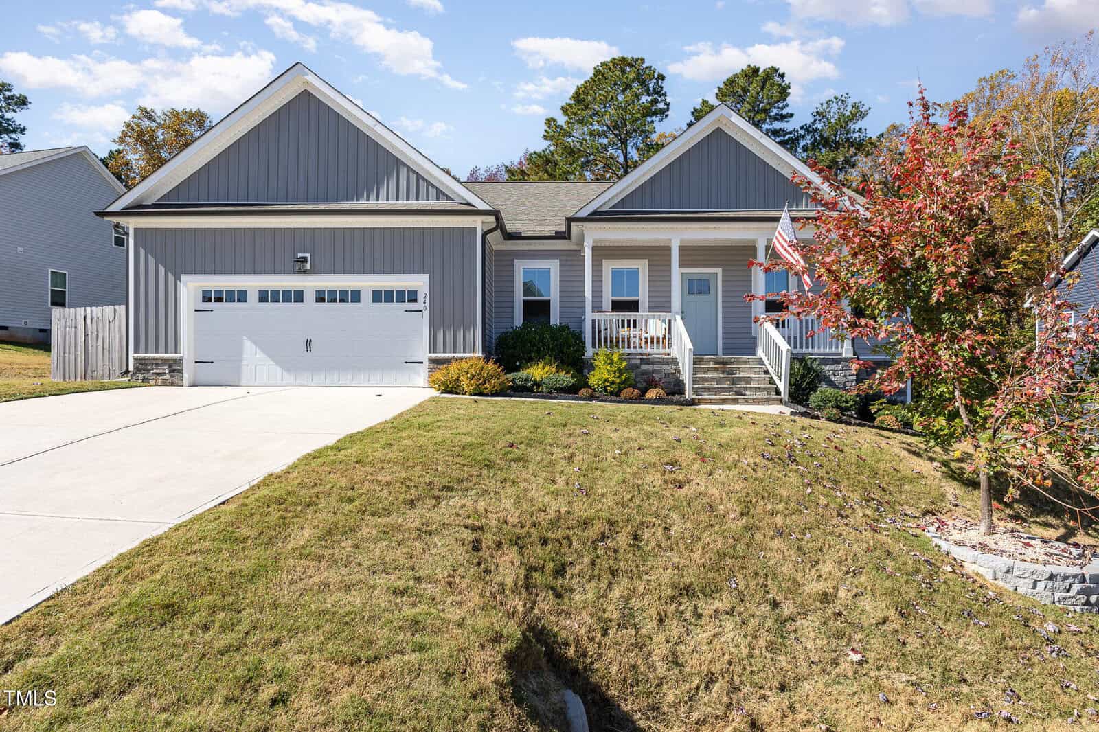 A modern gray house with white trim, attached two-car garage, front porch, landscaped yard, American flag, and steps leading to the front door. Trees and blue sky in the background.