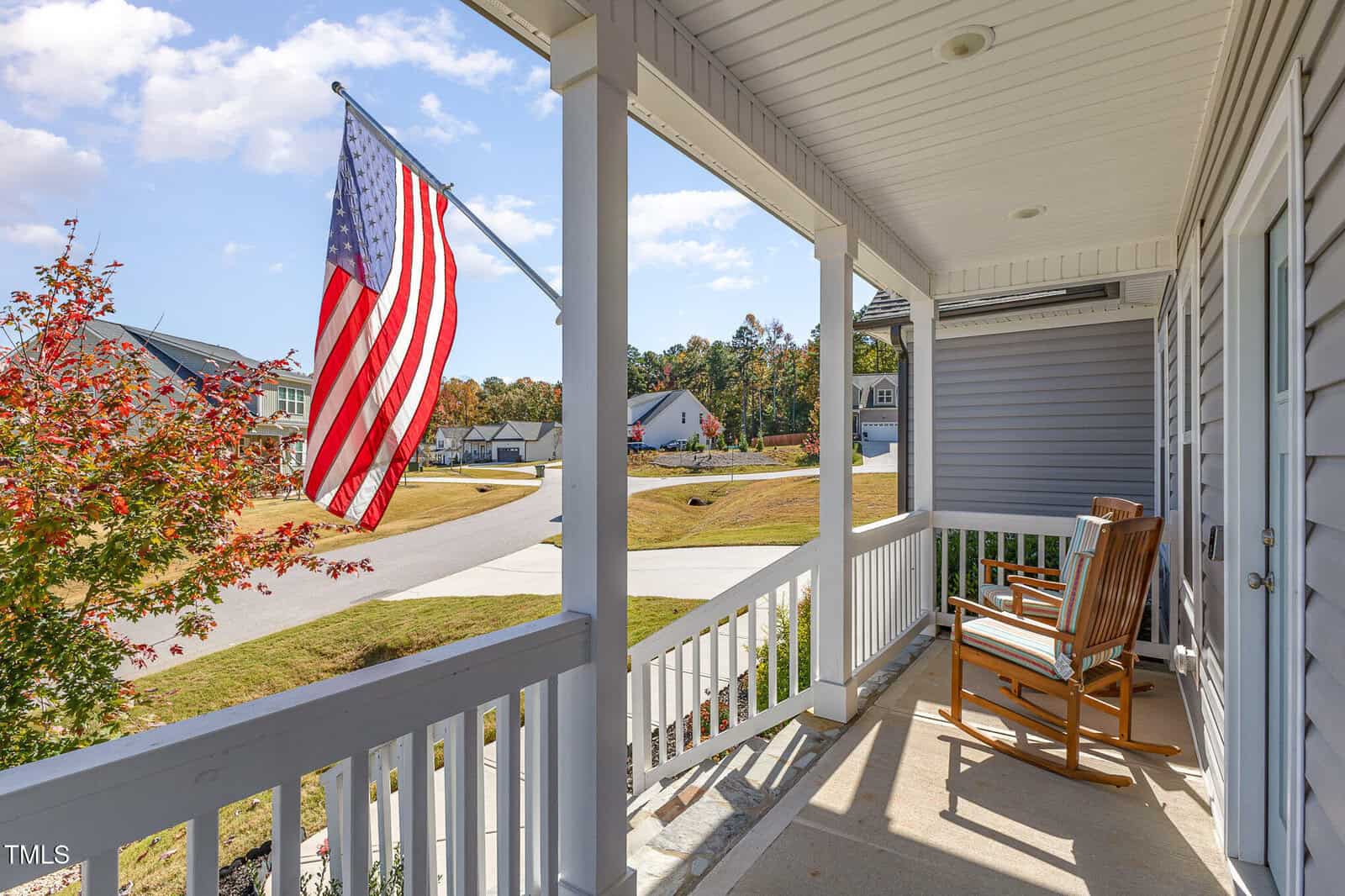 A front porch with two wooden rocking chairs and an American flag mounted on the railing, overlooking a suburban street with houses, trees, and a clear sky in the background.