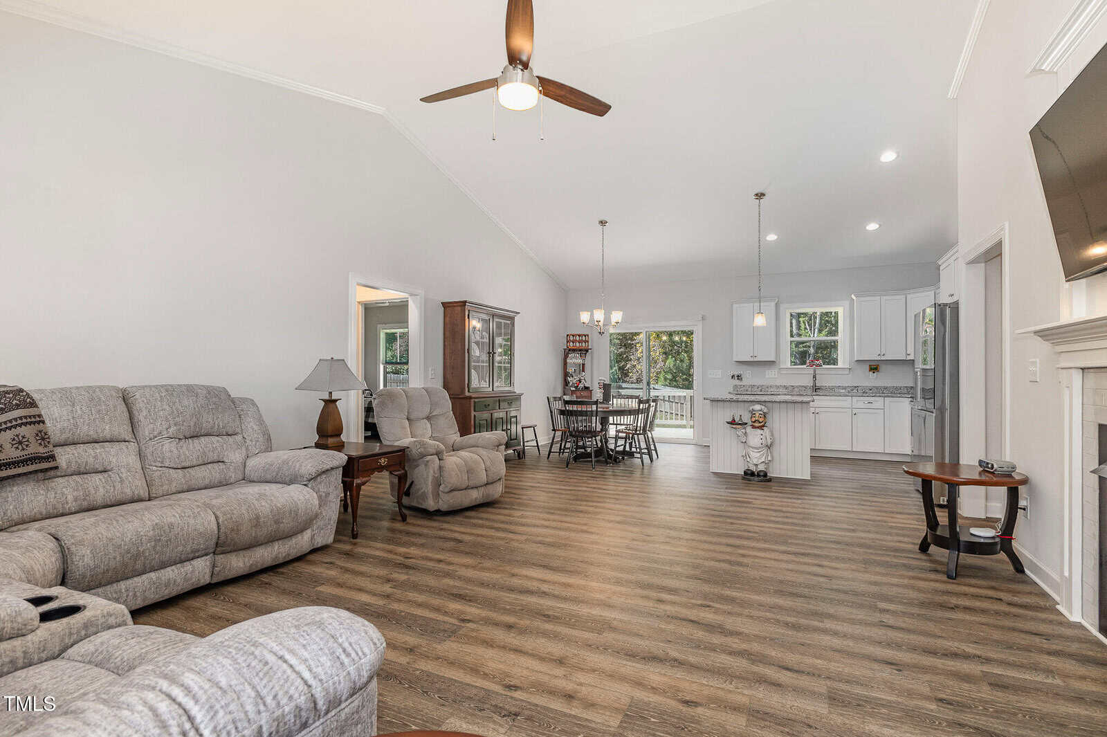 Spacious open-concept living room and kitchen with light wood flooring, gray recliner sofas, ceiling fan, dining area, white cabinets, center island, and large windows letting in natural light.