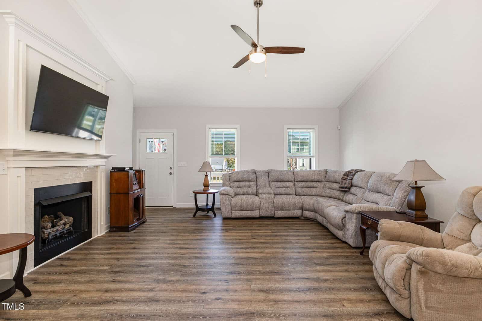 Spacious living room with wood flooring, large gray sectional sofa, beige recliner, two side tables with lamps, wall-mounted TV above a fireplace, and three windows letting in natural light.