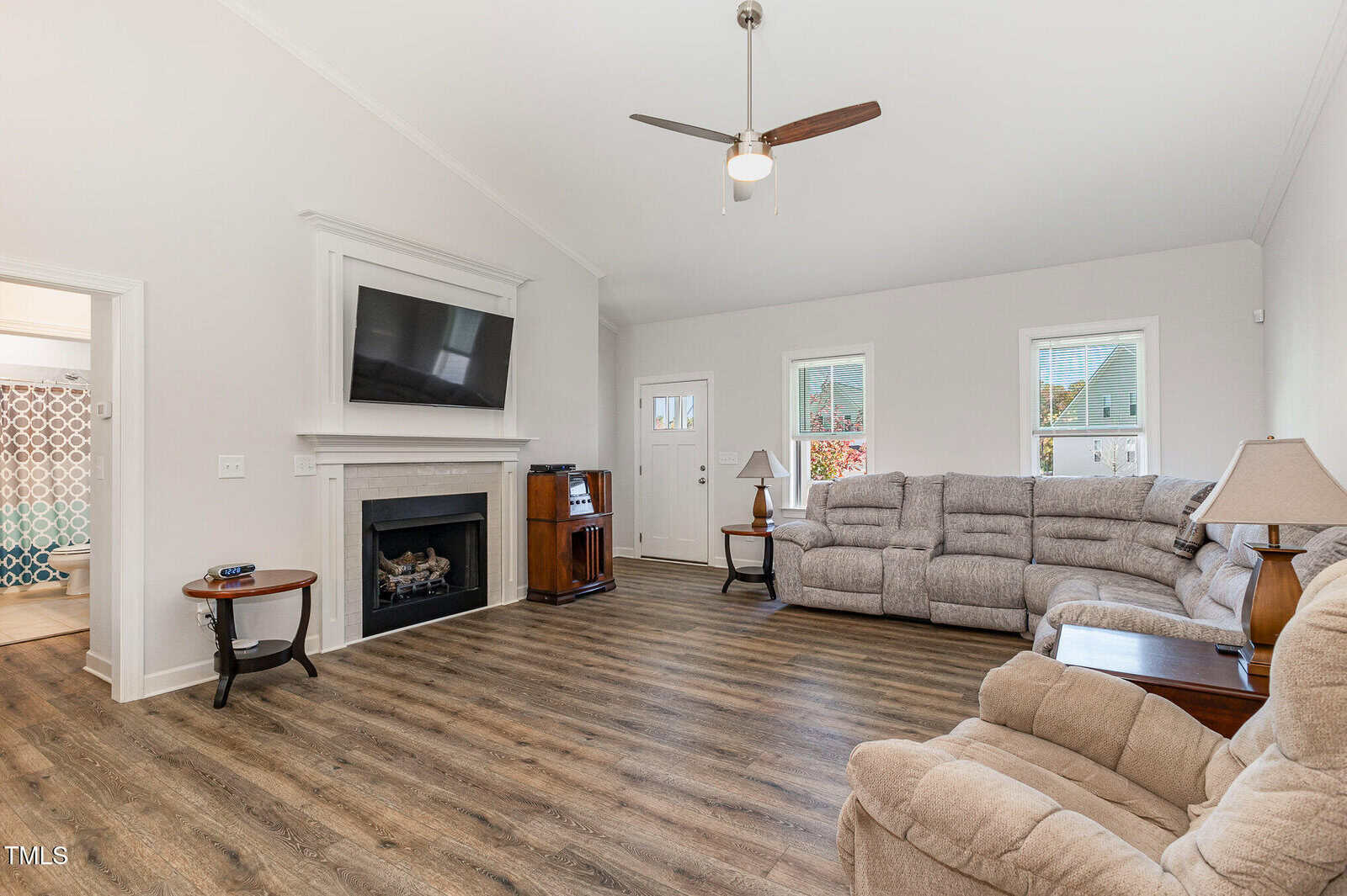 A bright living room with wood flooring, beige sectional sofas, a ceiling fan, a TV above a fireplace, a small round side table, and two lamps; three windows let in natural light.