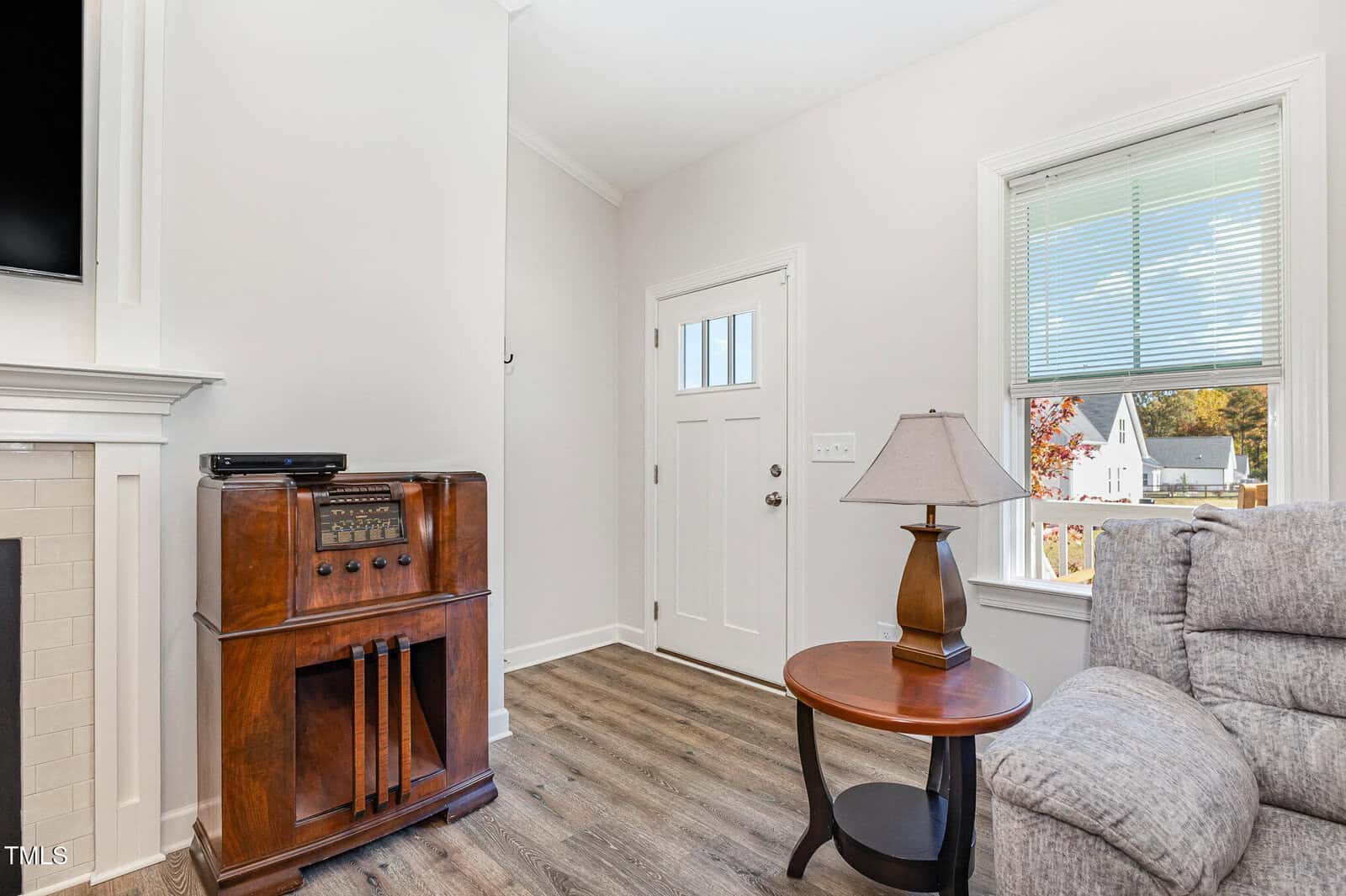 A cozy living room corner with a vintage radio console, round side table with a lamp, gray sofa, white door, and window with blinds letting in sunlight.