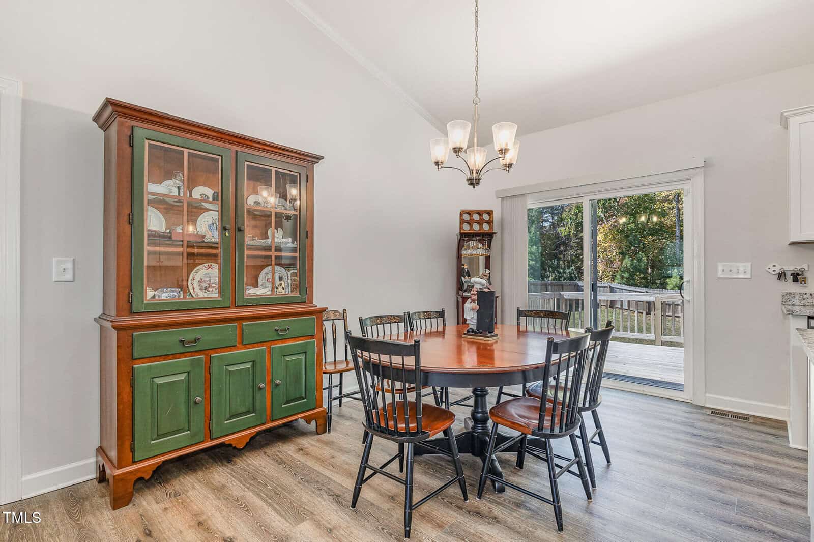 A dining area with a round wooden table, six black chairs, a china cabinet, grandfather clock, chandelier, and sliding glass door leading to a deck with trees visible outside.