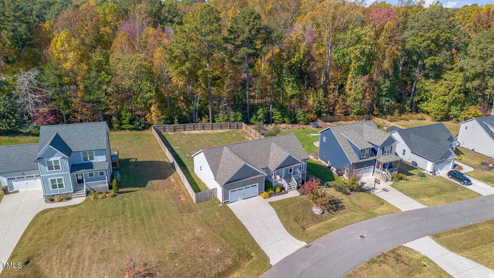 Aerial view of suburban homes with driveways, front lawns, and fenced backyards, bordered by a dense forest with autumn-colored trees. The street curves in front of the houses, and cars are parked in some driveways.