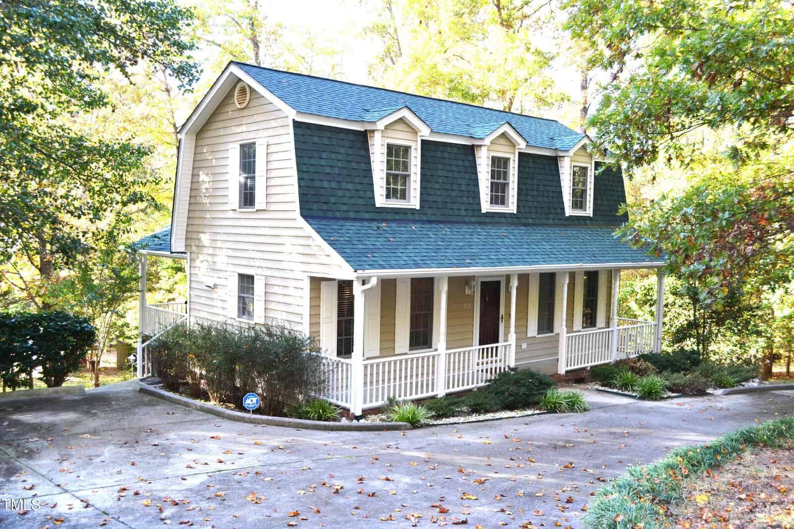 A two-story beige house with dark green roof shingles and white trim, featuring a covered front porch with railings and dormer windows, surrounded by trees and bushes on a sloped driveway.