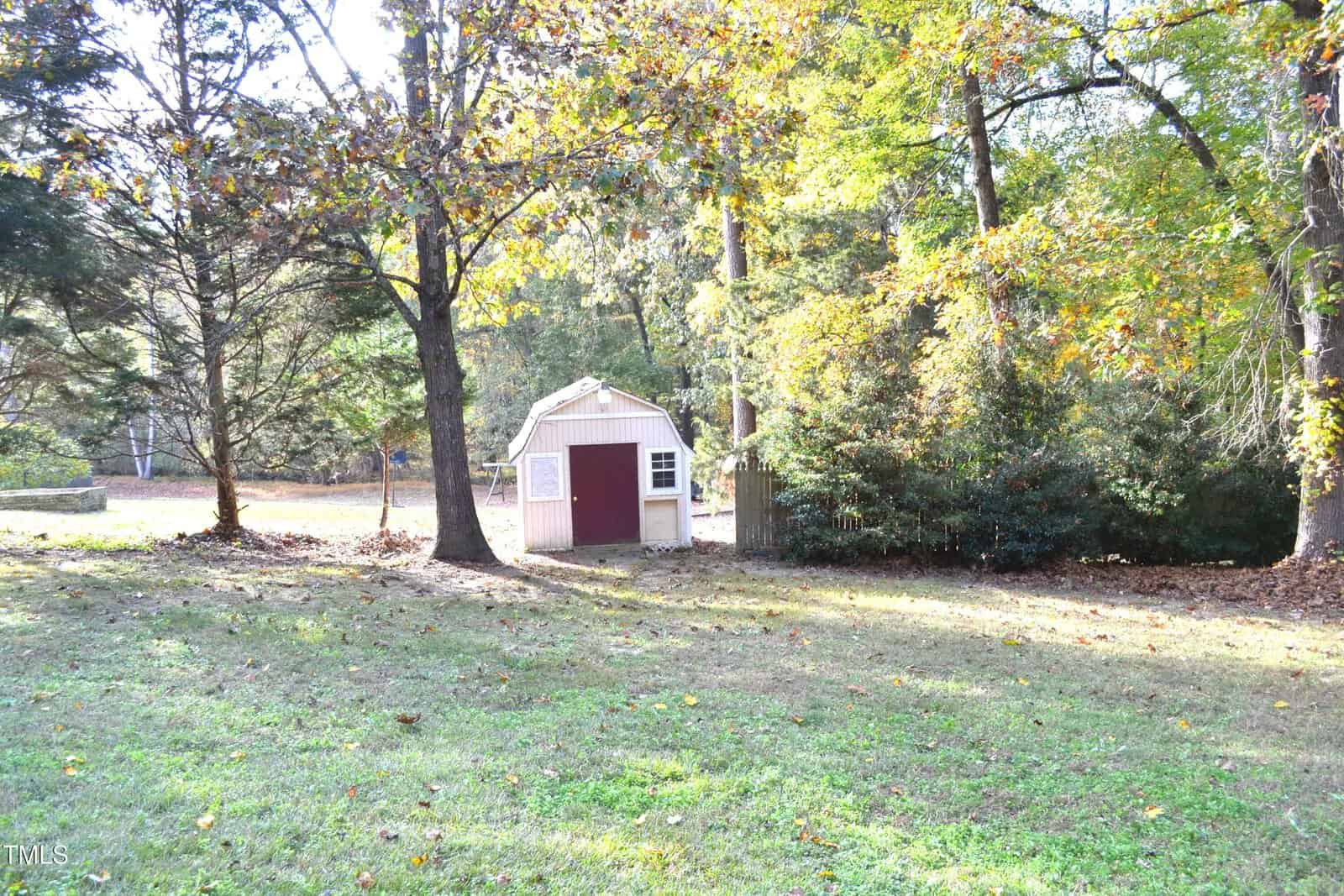 A small pink and white shed with a slanted roof sits among trees in a grassy backyard. Sunlight filters through the leaves, casting scattered shadows on the ground.