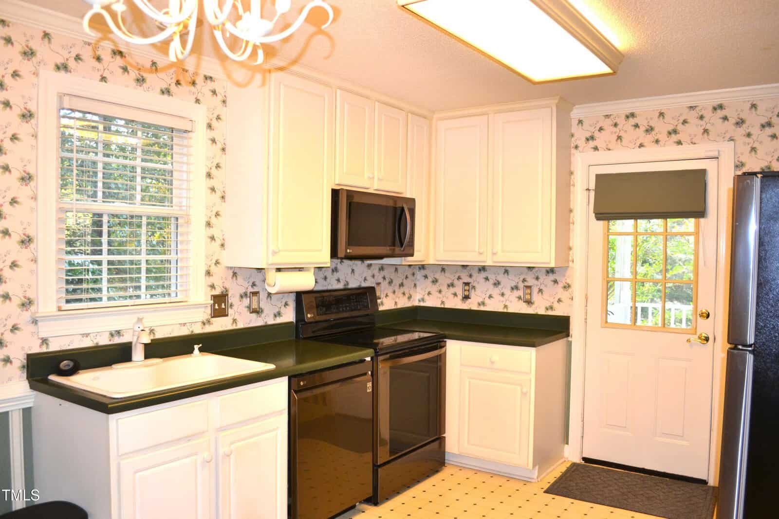 A kitchen with white cabinets, dark countertops, black appliances, a window above the sink, floral wallpaper, a door with a window, and a patterned linoleum floor. A chandelier and fluorescent light are on the ceiling.