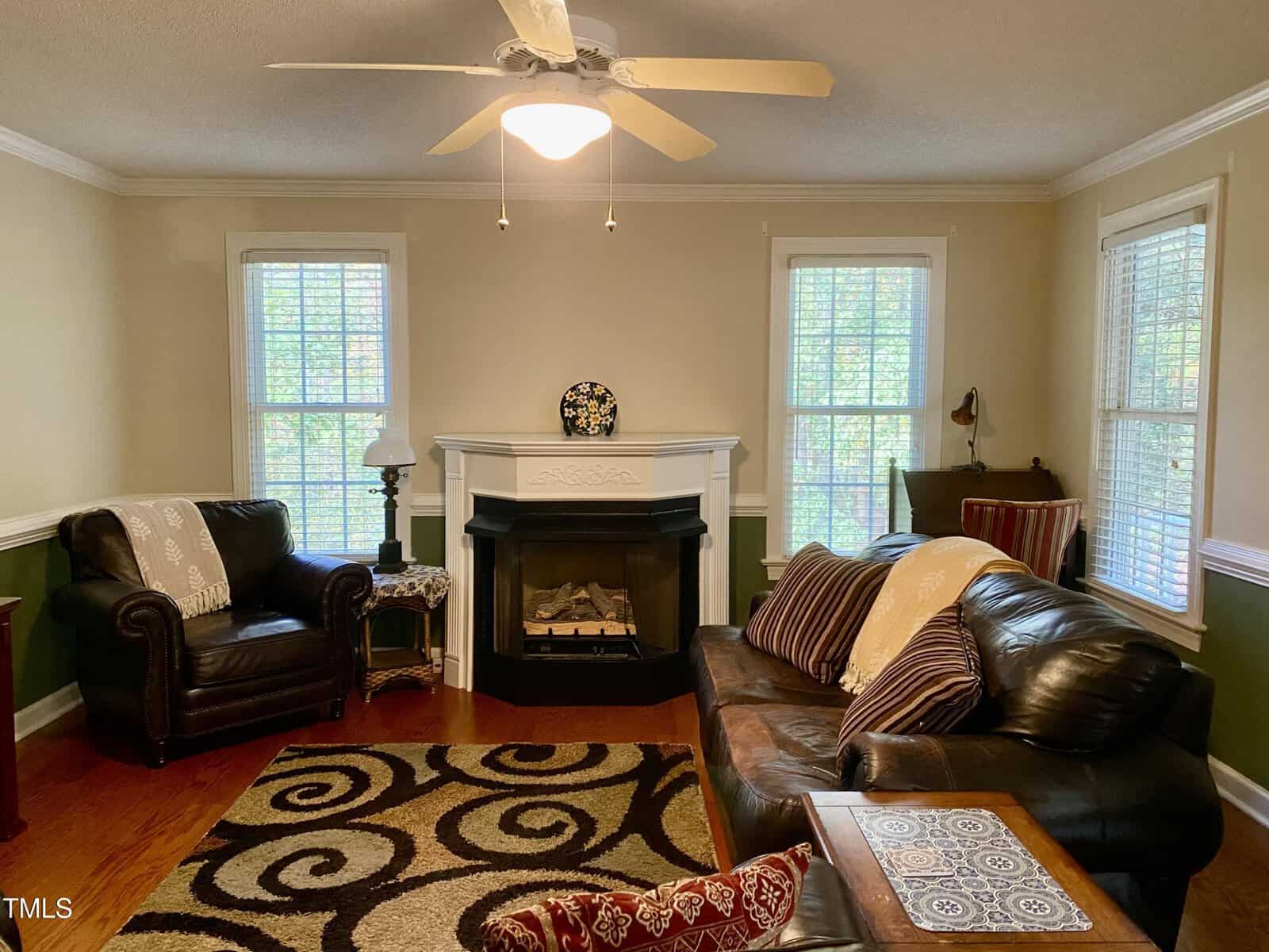 Cozy living room with brown leather sofa, armchair, patterned rug, fireplace, ceiling fan, and three windows with white blinds letting in natural light. Pillows and throws add comfort to the space.