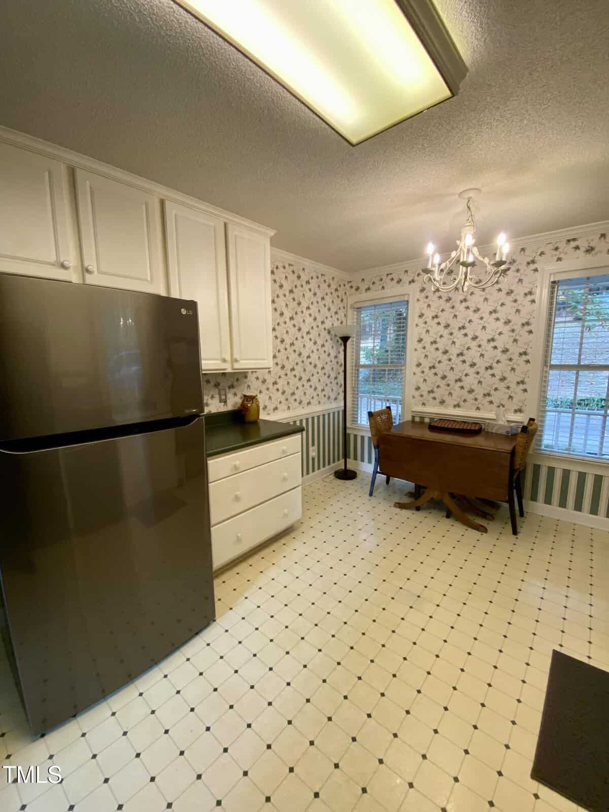 A kitchen with white cabinets, a stainless steel refrigerator, floral wallpaper, a small dining table with two chairs, large windows, and a chandelier.