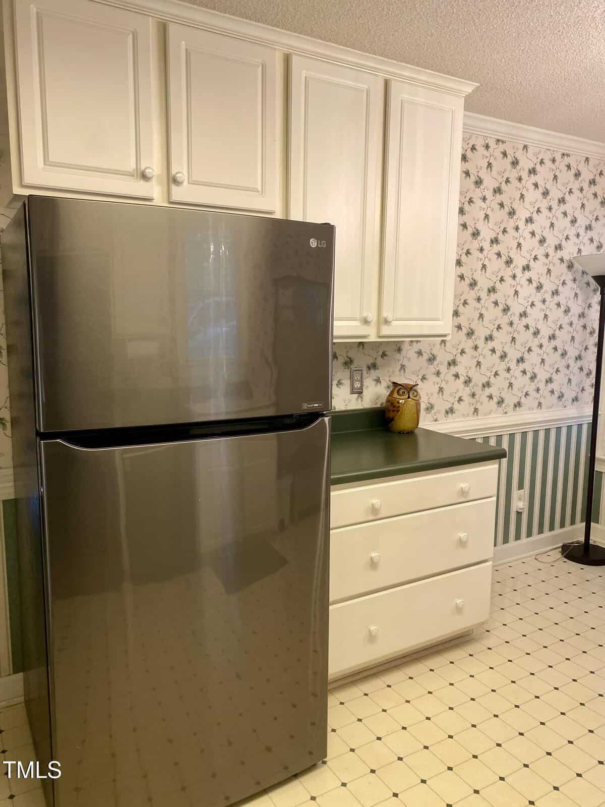 A stainless steel refrigerator next to white cabinets and drawers with green countertops; a ceramic owl container sits on the counter. The wall has floral wallpaper and there is a floor lamp in the corner.